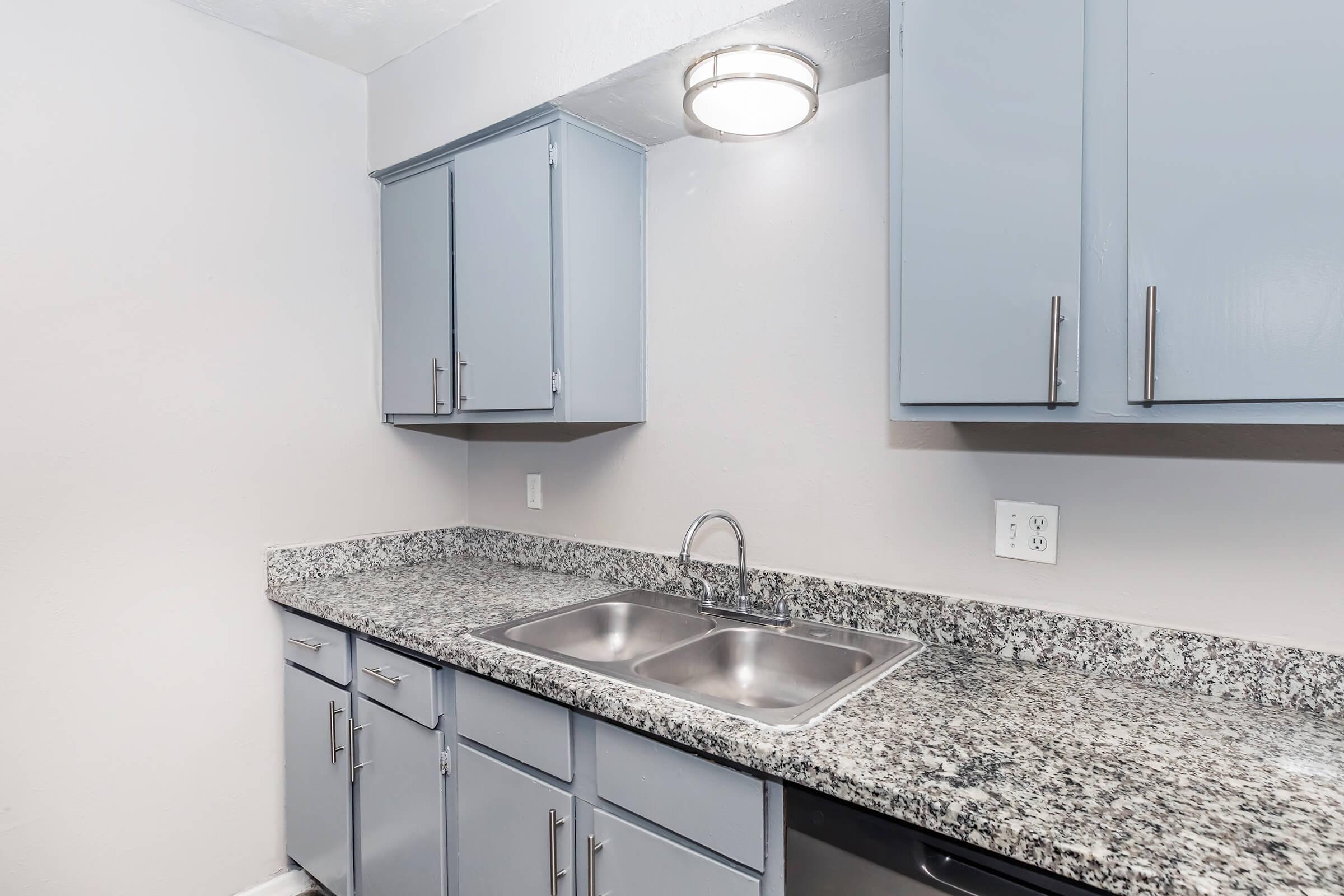 A modern kitchen featuring light blue cabinets with silver handles, a double stainless steel sink, and a granite countertop. The walls are painted a light gray, and there is a ceiling light fixture above. The overall design is clean and contemporary, with appliances and a tidy appearance.