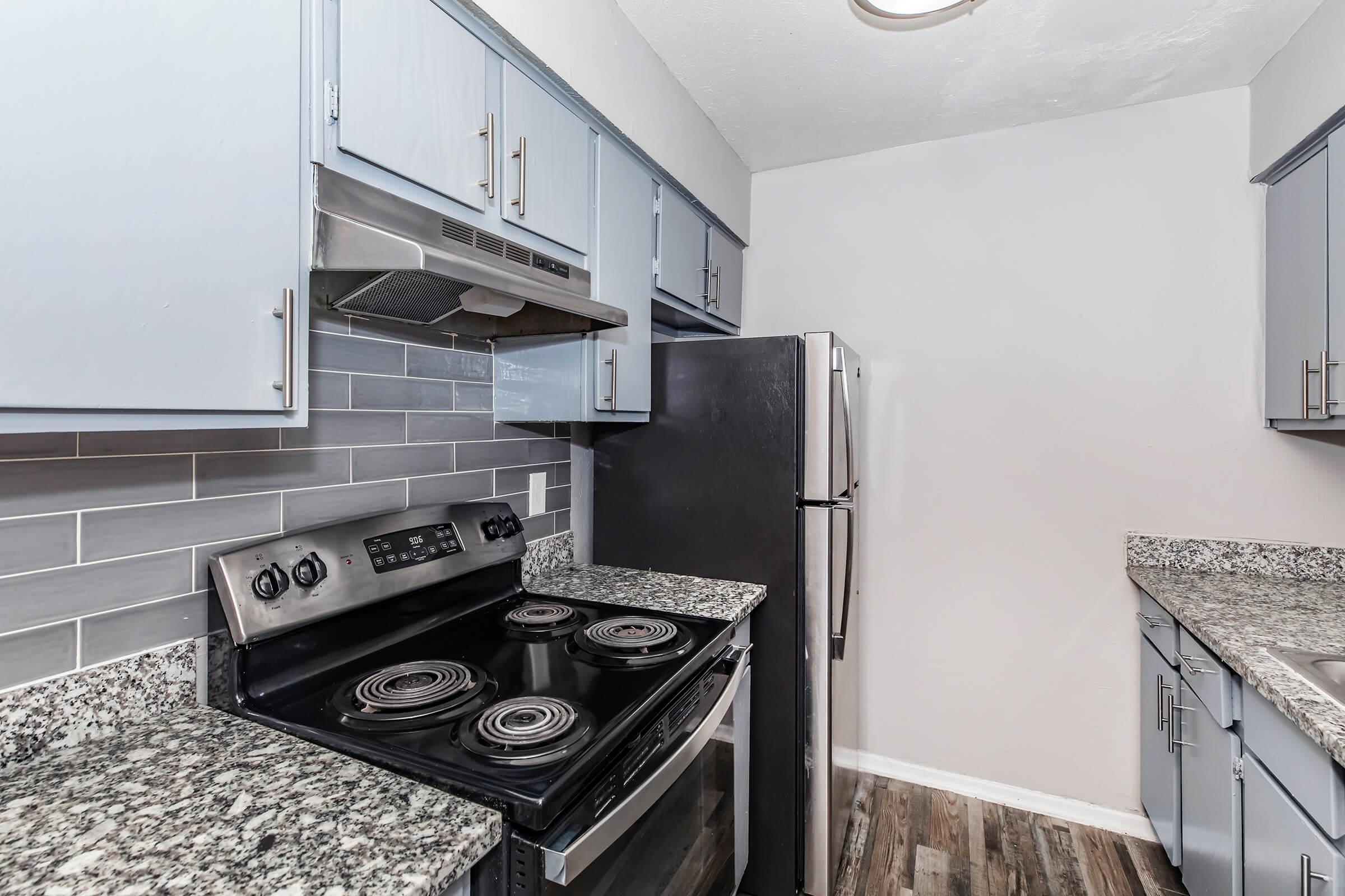 A modern kitchen featuring gray cabinets, a stainless steel refrigerator, and a black electric stove. The backsplash is made of gray tiles, and the countertops have a speckled granite design. Soft lighting illuminates the space, showcasing a clean and stylish cooking area.