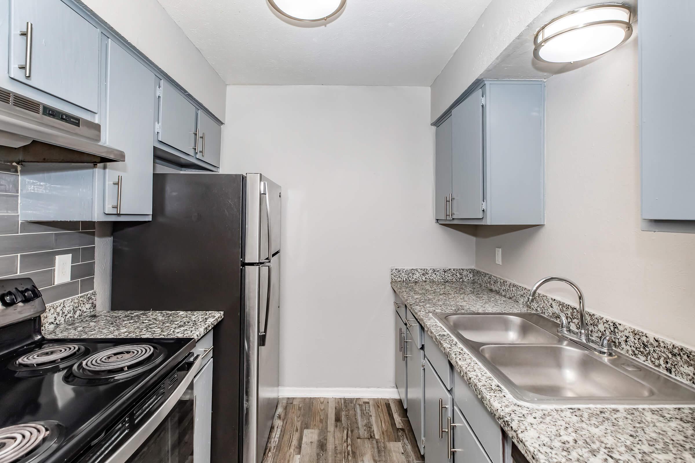 A modern kitchen featuring light blue cabinets, a stainless steel refrigerator, an oven with a black cooktop, and a double sink. The countertops are a speckled gray granite, and the space has laminate flooring. Bright overhead lighting enhances the clean, contemporary design.