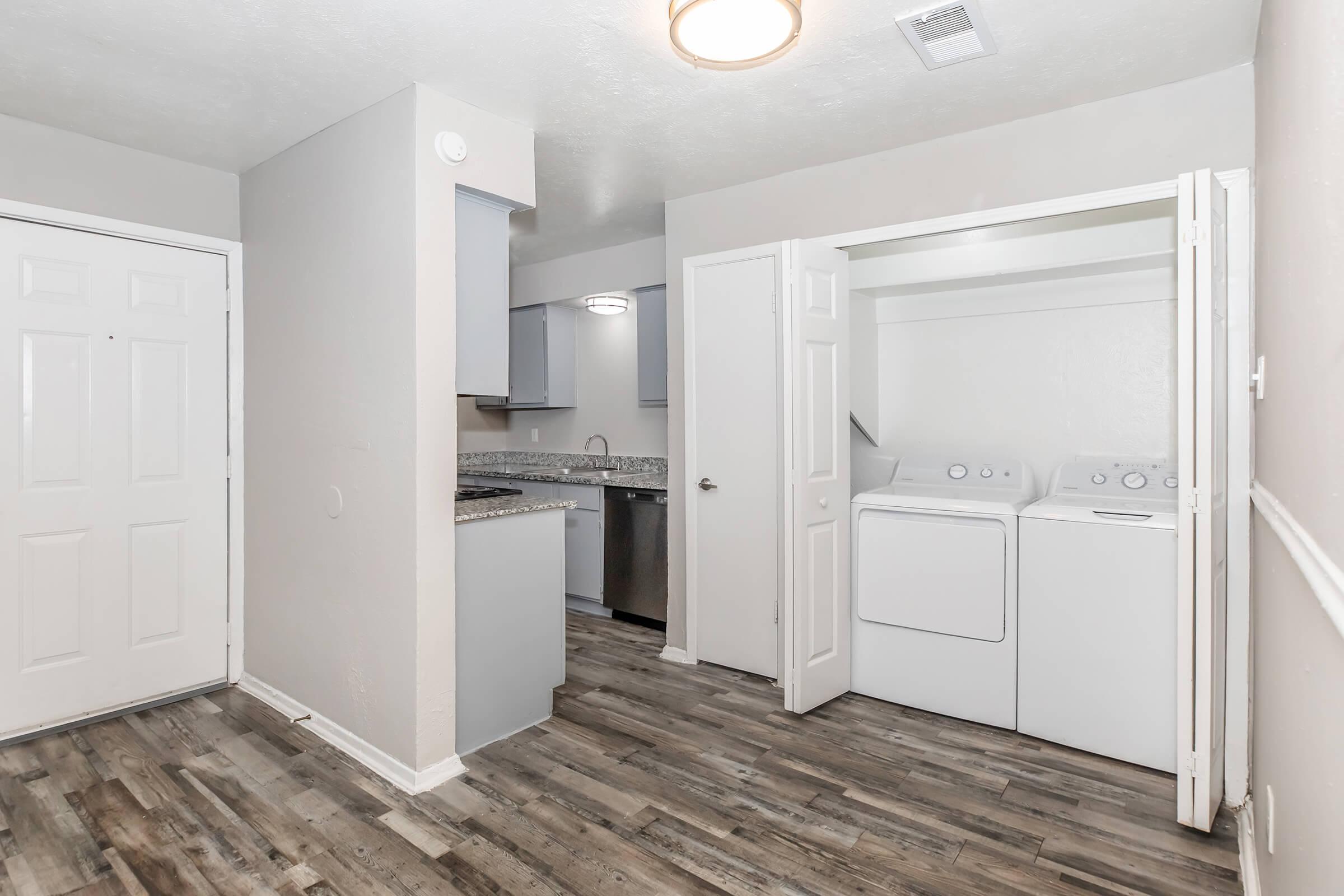 An interior view of a laundry area in an apartment, featuring a set of white washing machines and dryers. The space has light-colored walls, wooden flooring, and a kitchen area visible in the background with gray cabinets and granite countertops. Natural light illuminates the room.