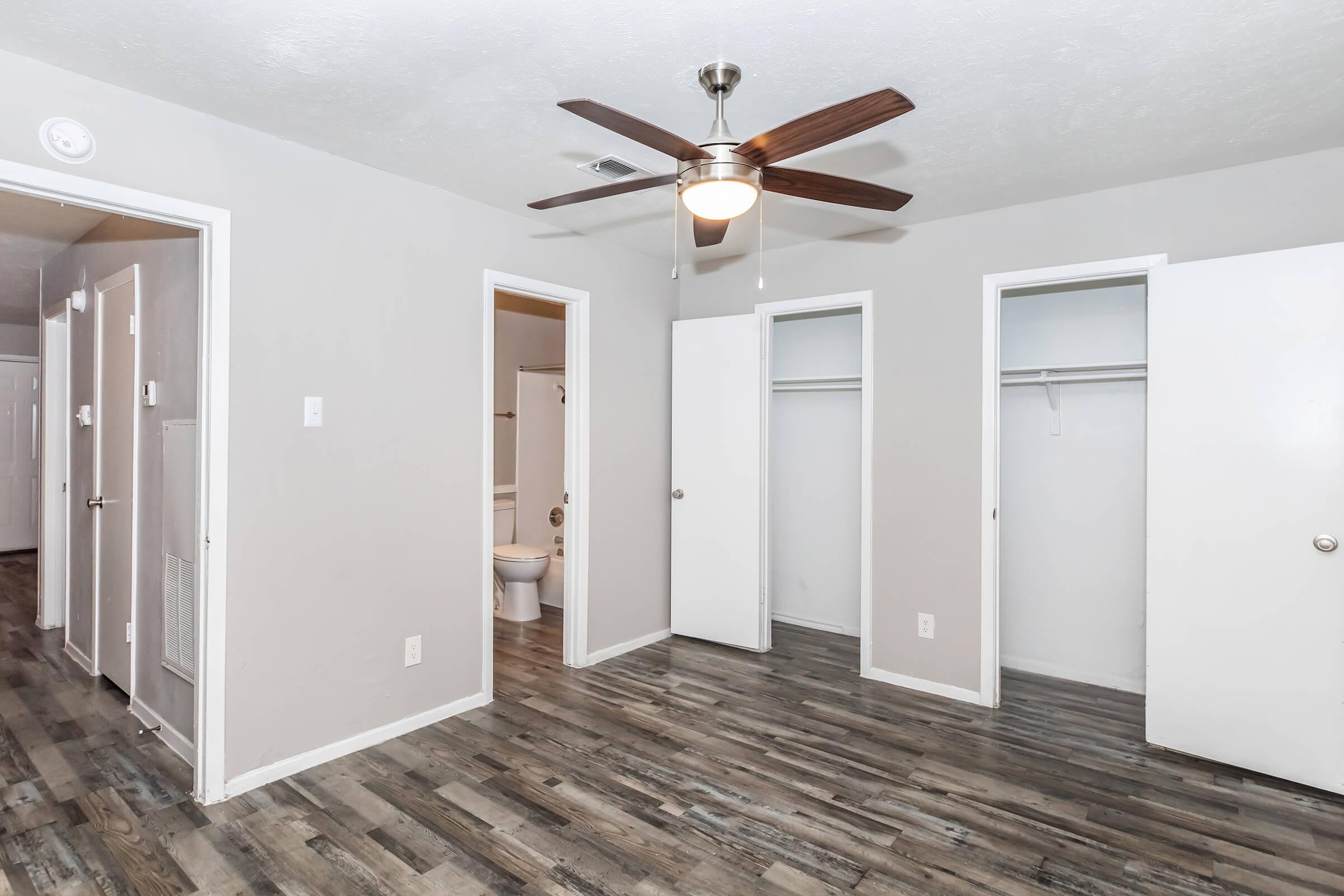 A spacious, well-lit room featuring a ceiling fan and laminate flooring. Two closets are on the right side, and an open doorway leads to a bathroom. The walls are painted a light gray, and the room has a neutral and modern appearance.