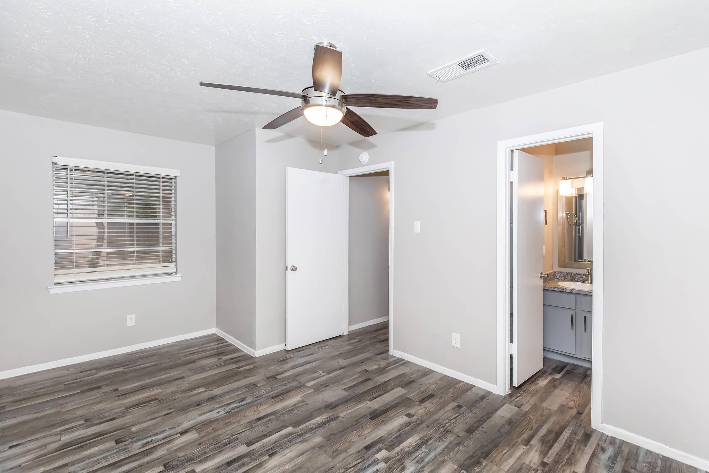A well-lit interior of a room featuring a ceiling fan, hardwood-look flooring, and light gray walls. There is a window with blinds on the left, a closed white door in the center leading to another space, and an open door on the right leading to a bathroom with a mirror and sink.