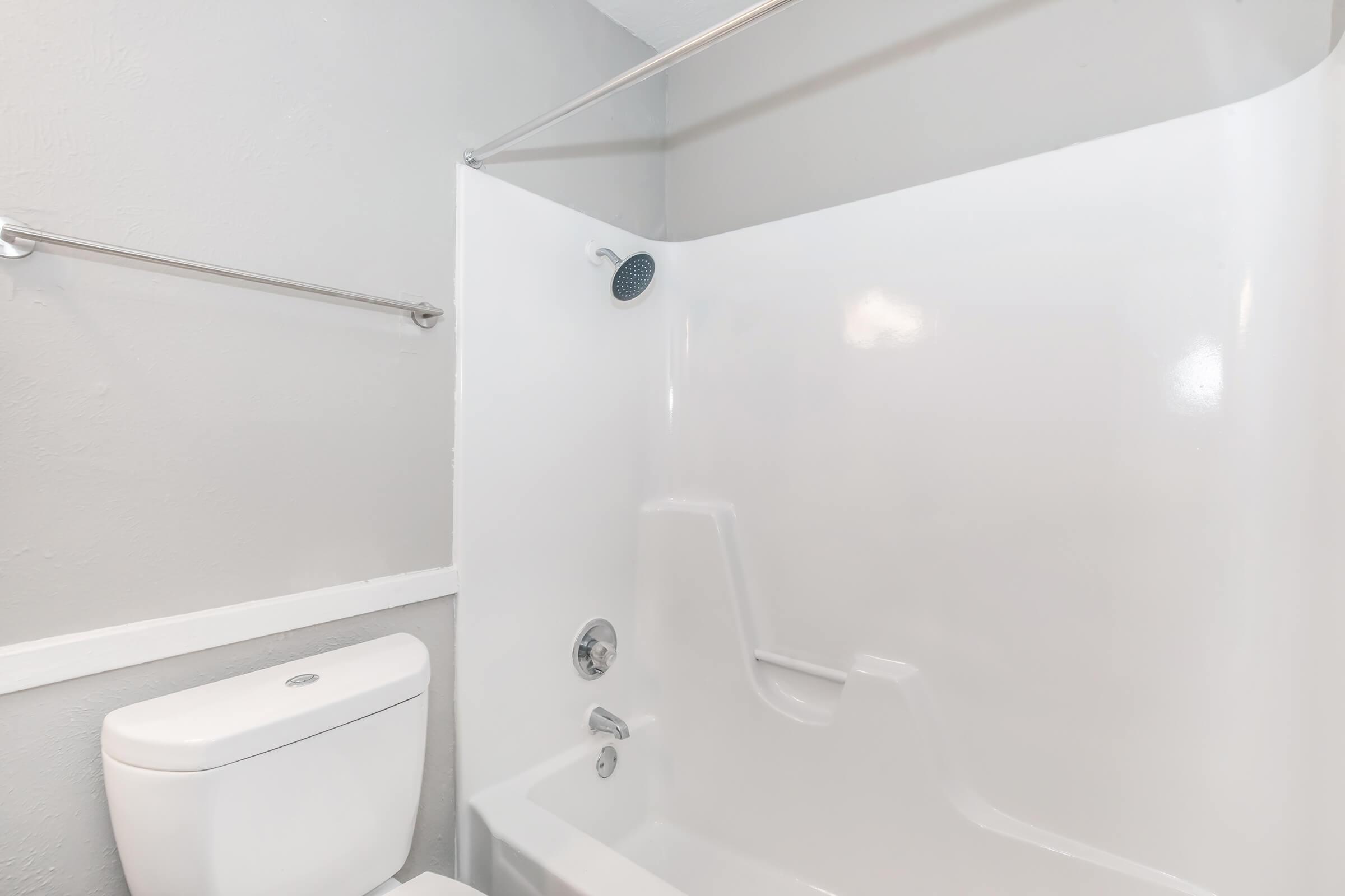 A clean and modern bathroom featuring a white bathtub with a shower, a wall-mounted showerhead, and a sleek toilet. The walls are painted in a light gray shade, creating a fresh and minimalist aesthetic.