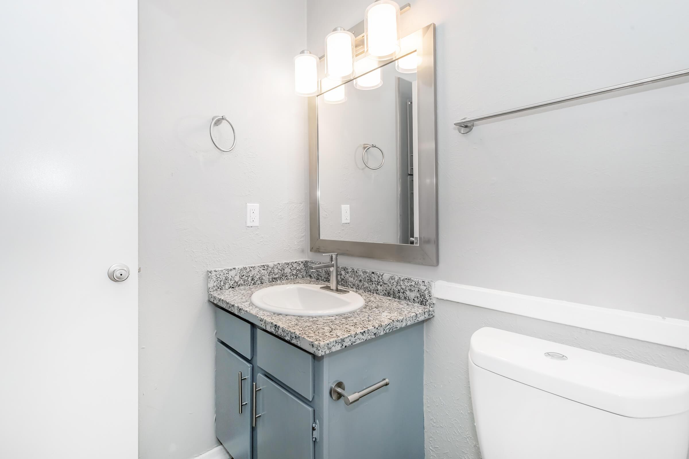 A modern bathroom featuring a granite countertop with a circular sink, a sleek mirror above, and three pendant lights. The cabinetry is painted in a soft blue, and there is a white toilet to the right. The walls are painted in a light gray color, and a towel rack is mounted above the countertop.