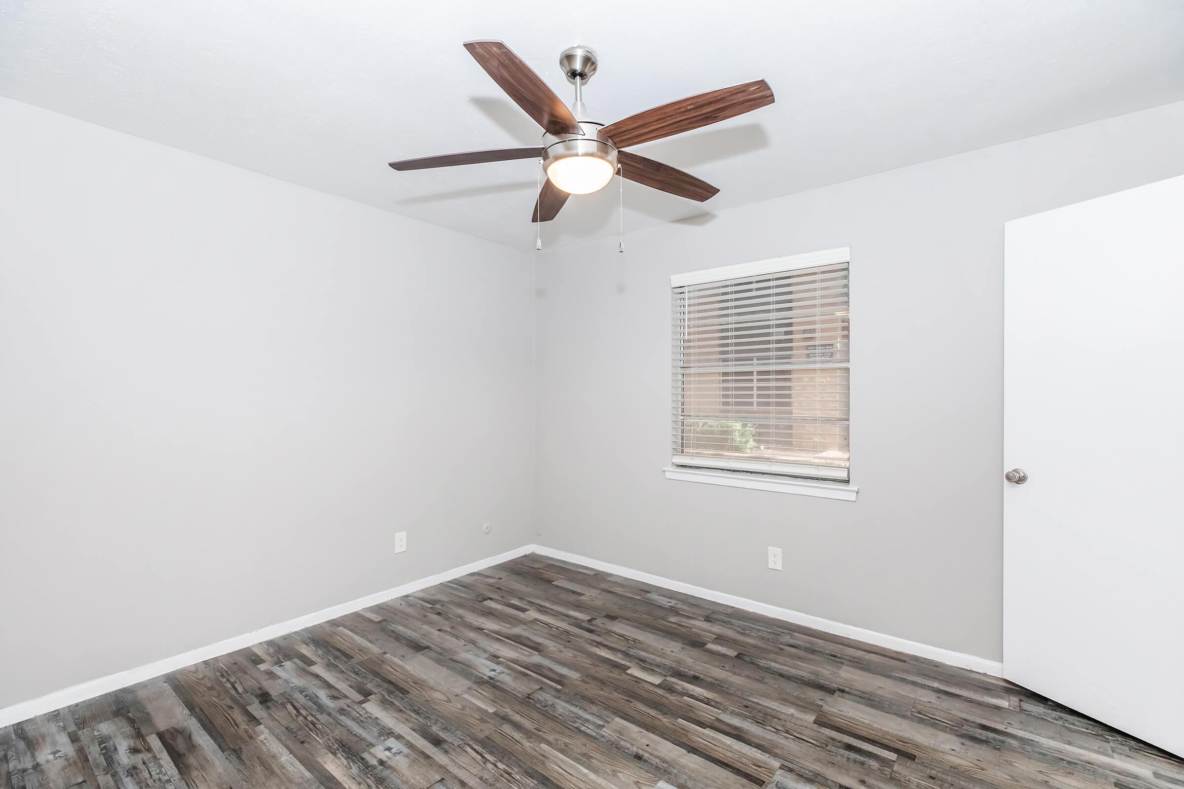 Empty room with light gray walls and a wooden floor. A ceiling fan with wooden blades hangs above. There is a window with blinds allowing natural light, and a white door is visible on the right side of the image. The space appears clean and uncluttered, ideal for various uses.