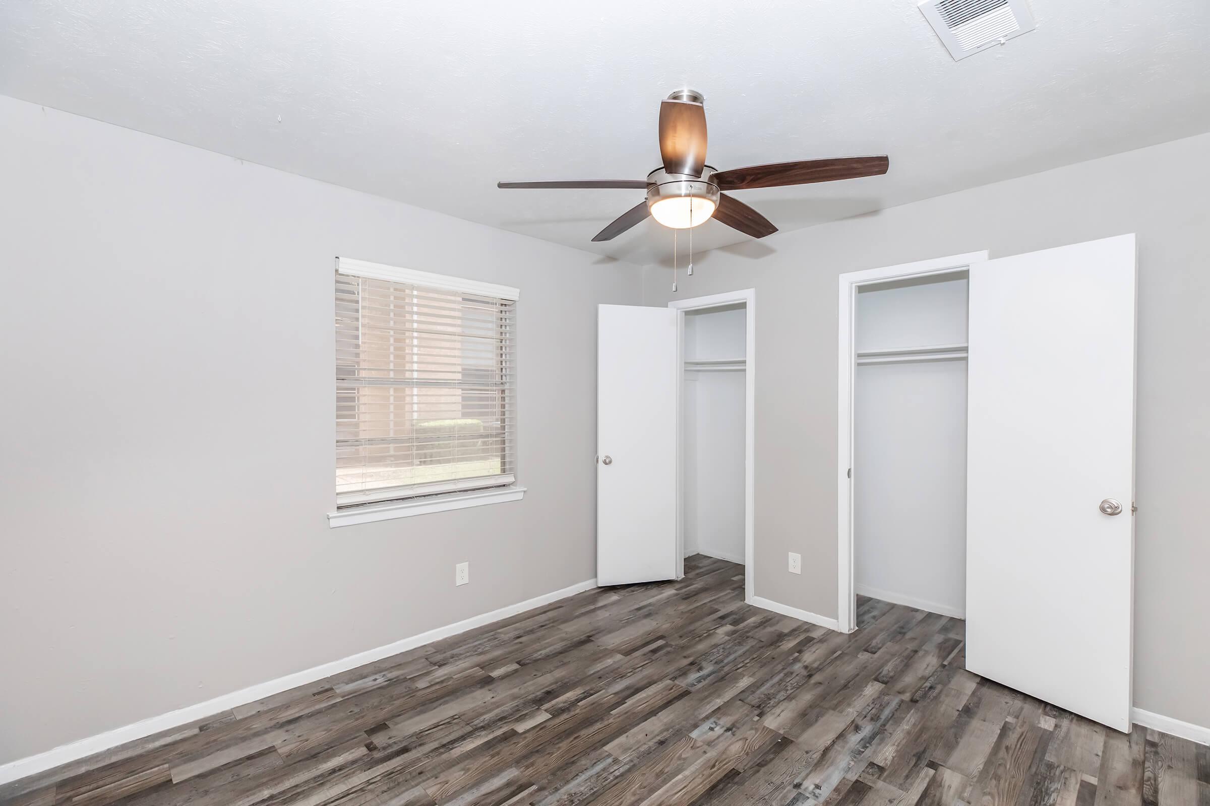 A small room with light gray walls and a ceiling fan. The floor is made of wood-like laminate. There are two white closet doors on opposite walls, both slightly open, showing empty closet spaces. A window with blinds allows natural light to enter the room.