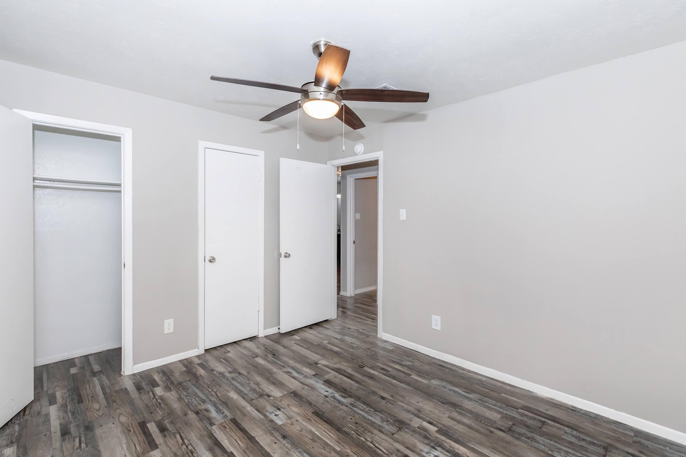 A well-lit room featuring light gray walls and a ceiling fan with wooden blades. There are two closed white doors on the left, leading to a closet and another room. The floor is made of textured wood, adding warmth to the space. Natural light illuminates the room, creating a cozy atmosphere.