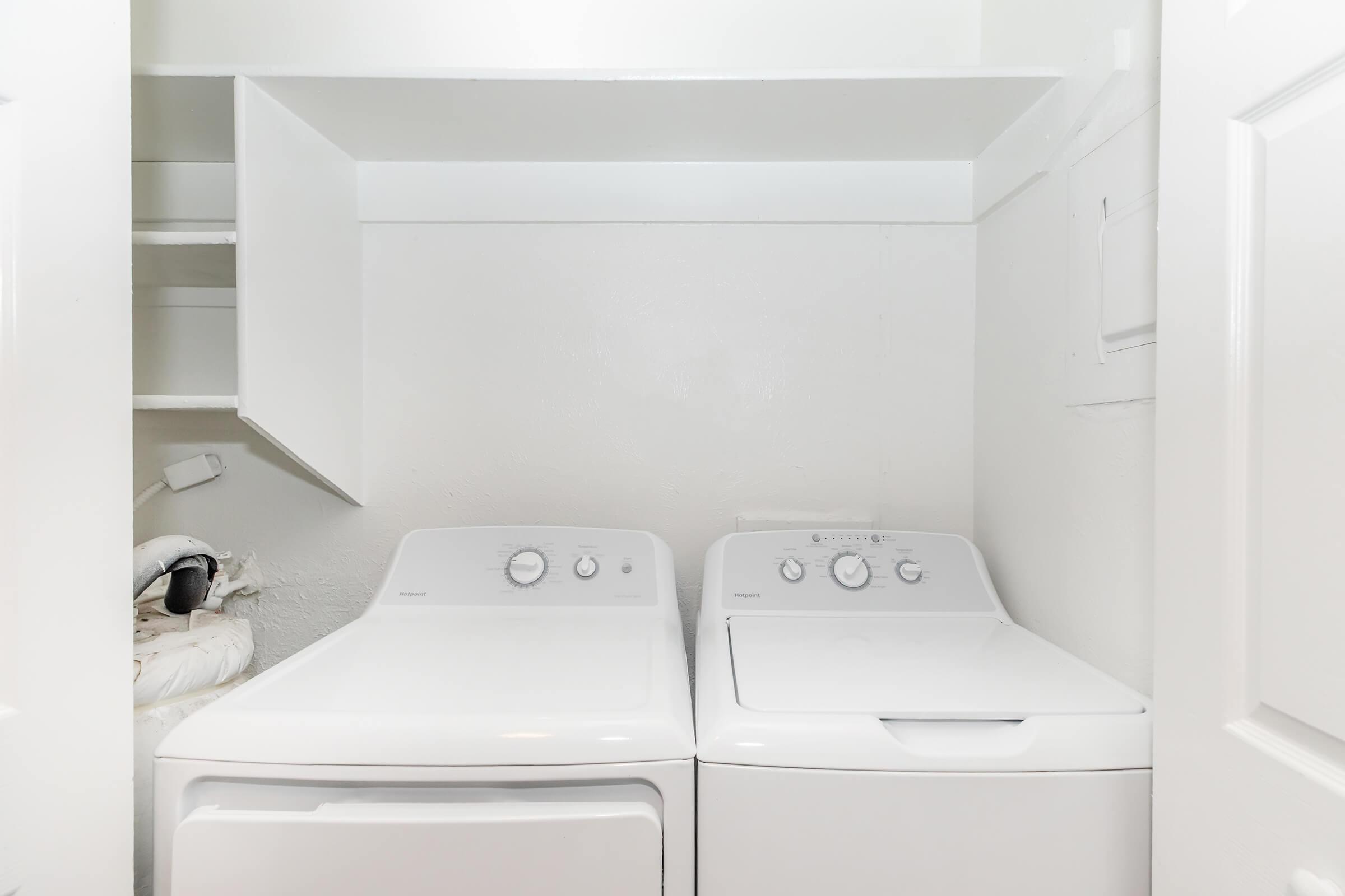 A laundry room featuring a stacked white washer and dryer. The wall is painted white, and there is a shelf above the appliances. The space is clean and minimalist, with light reflecting off the surfaces.