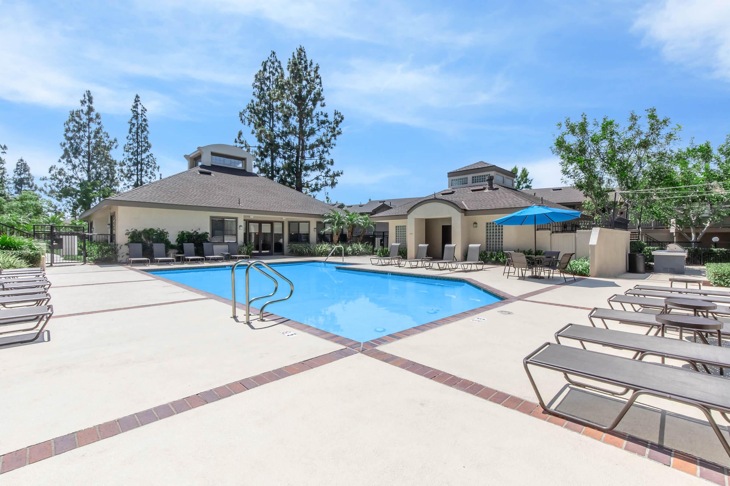 A bright outdoor swimming pool surrounded by lounge chairs and shaded areas. In the background, there are trees and buildings. A large blue umbrella provides shade near the poolside. The area is well-maintained and sunny, ideal for relaxation and leisure activities.
