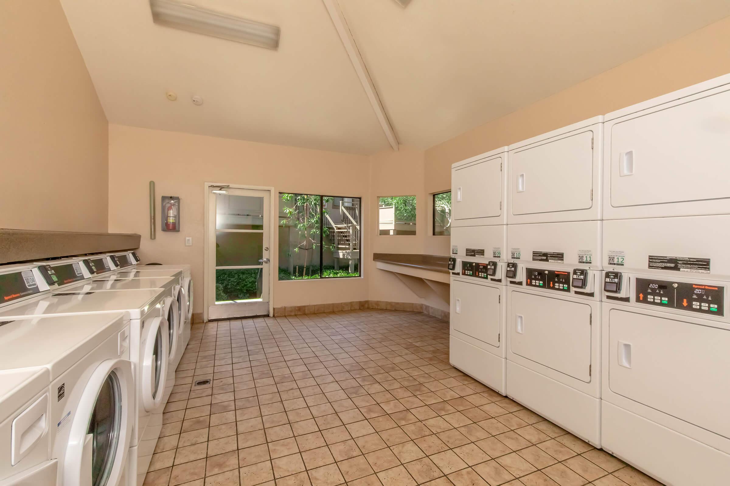 A well-lit laundry room featuring multiple washing machines and dryers lined against the wall. There's a countertop for folding clothes and a door leading to an outdoor area. Large windows allow natural light to fill the space, enhancing the clean and organized atmosphere.