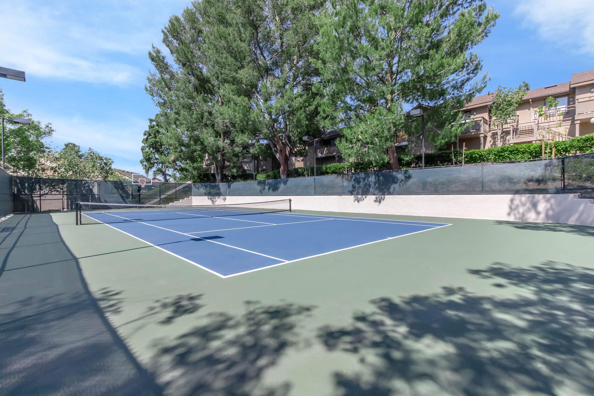 View of a tennis court featuring blue and green surfaces, surrounded by trees and residential buildings. The court is empty and well-maintained, with a clear blue sky overhead. Shadows from the trees are visible on the court, creating a peaceful outdoor atmosphere.