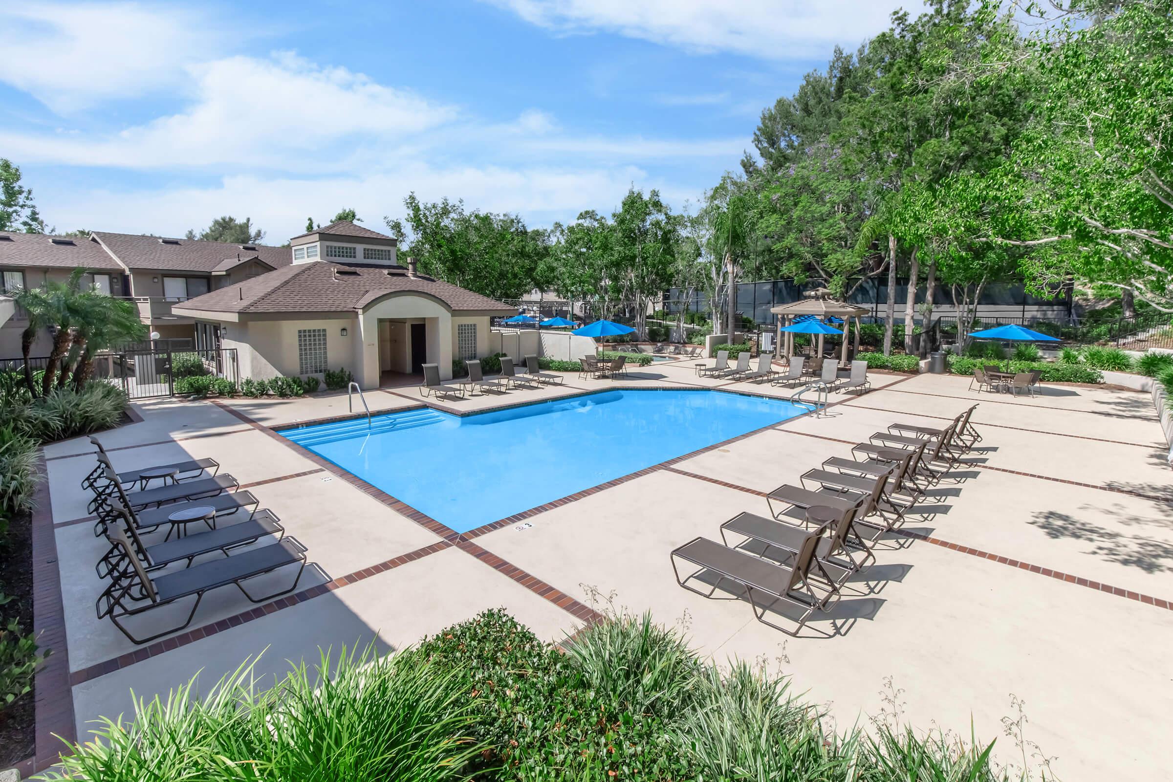 A serene outdoor swimming pool area surrounded by lounge chairs and umbrellas, set in a landscaped environment with trees. The pool features a light blue water surface, and the area is well-lit with clear skies and a pleasant atmosphere, ideal for relaxation and leisure activities.