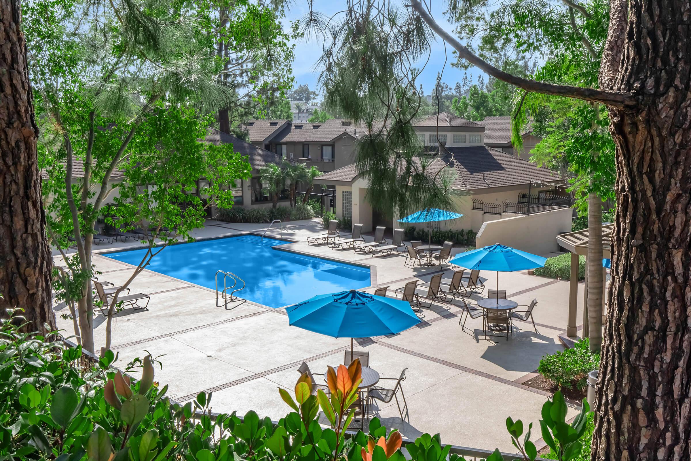 A serene outdoor pool area surrounded by lush greenery, featuring a large blue swimming pool, lounge chairs, and round tables with blue umbrellas. The scene is bright and inviting, set against a backdrop of residential buildings with a clear blue sky above.
