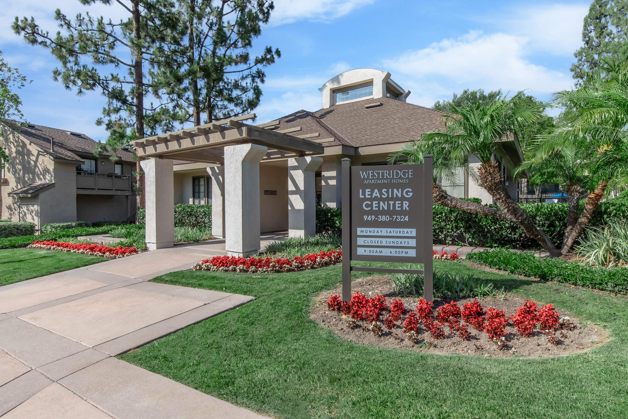 A well-maintained leasing center for Westridge, featuring a modern building design with a covered entrance. Surrounding the building are manicured lawns, vibrant flower beds in shades of red, and tall palm trees, set against a clear blue sky. The sign includes contact information for leasing inquiries.