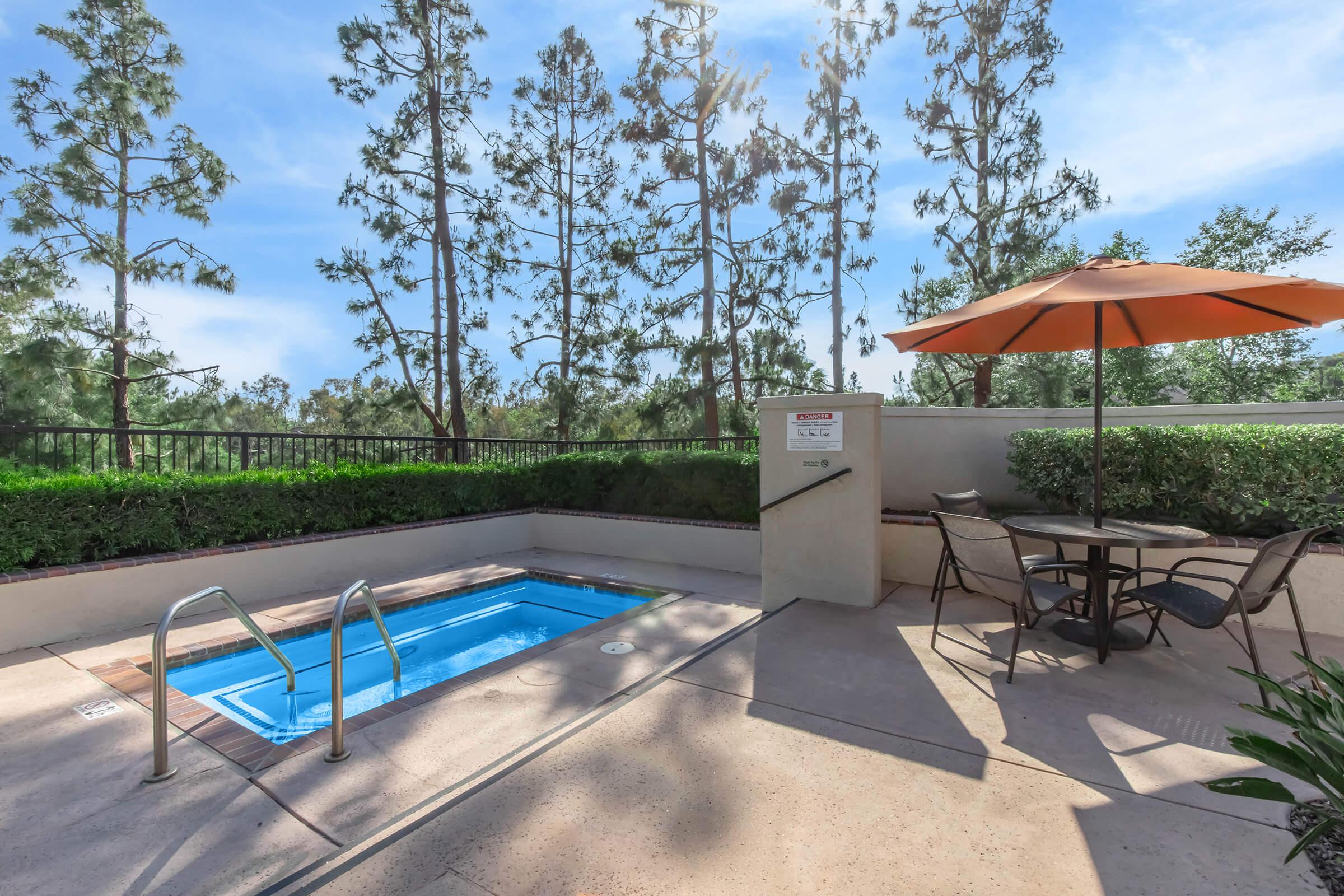 A small outdoor hot tub beside a patio area with a table and chairs. An orange umbrella provides shade. Lush green foliage and tall trees are visible in the background under a clear blue sky. The overall setting is tranquil and inviting, perfect for relaxation.