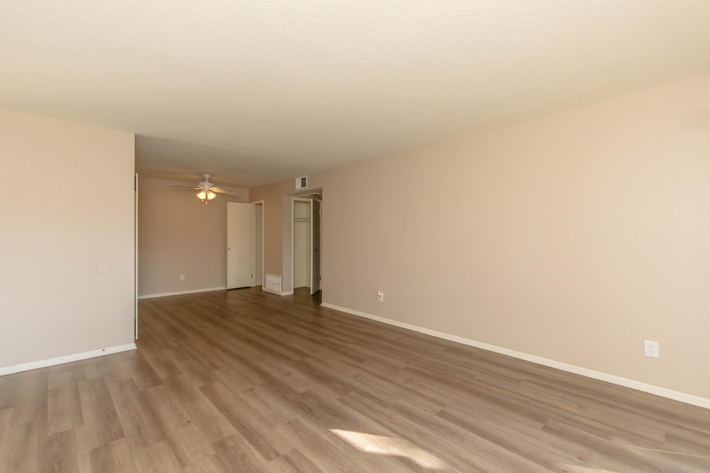 A spacious, empty room with light-colored walls and wood laminate flooring. A ceiling fan is visible, and there are two doorways leading to other areas. Natural light fills the space, creating a bright and airy atmosphere. Ideal for a living area or studio setup.