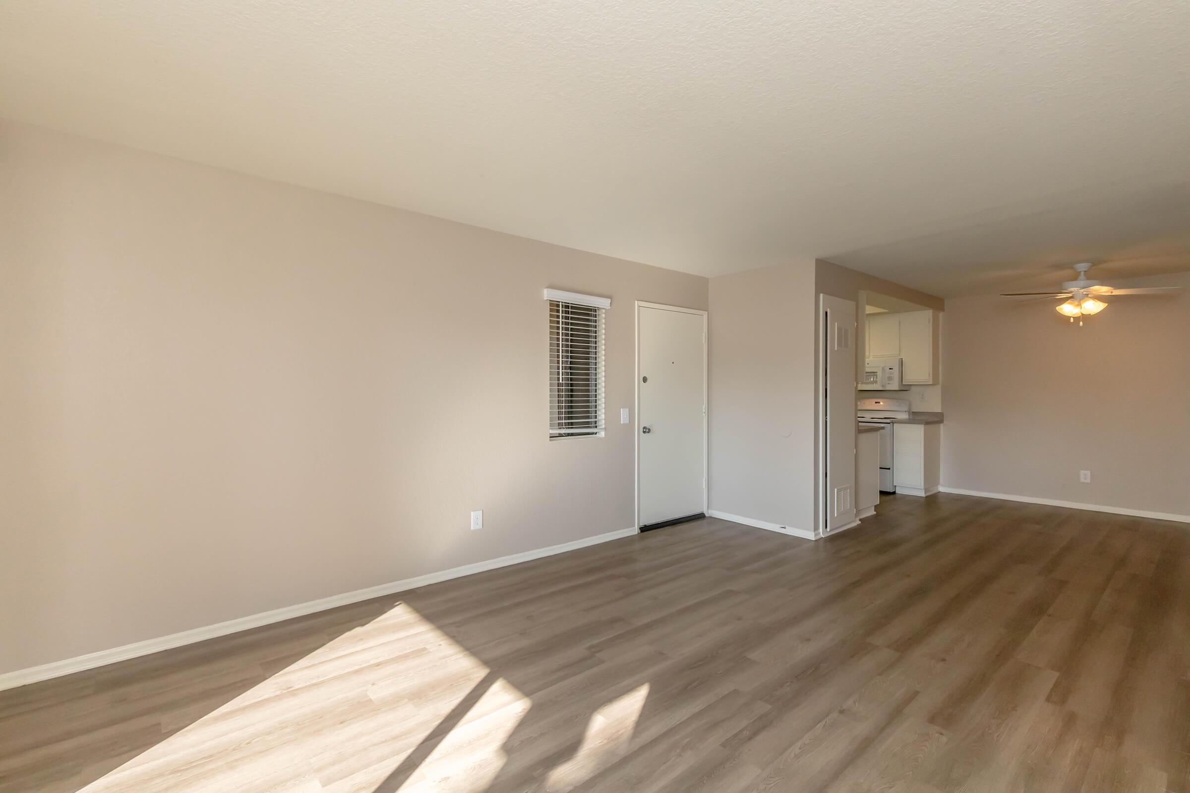 Spacious living area with light-colored walls, large windows, and vinyl flooring. The entrance door is visible on one side, while the kitchen area can be seen in the background. A ceiling fan is installed, and the overall layout is open and inviting.