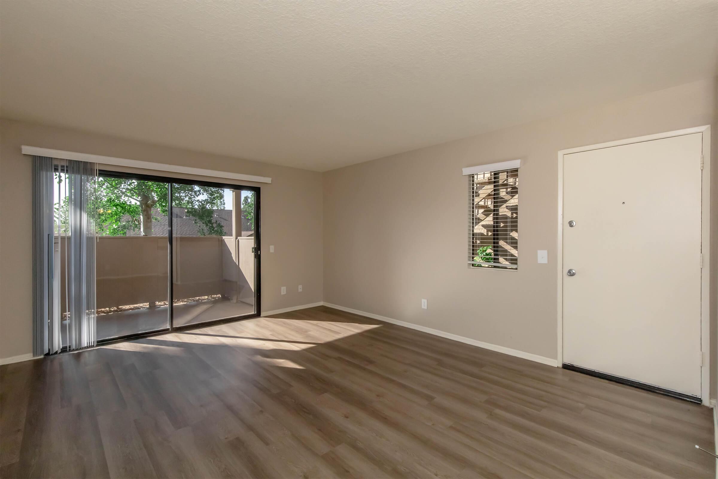 A spacious living room featuring light beige walls, large window with vertical blinds, and a sliding glass door leading to an outdoor area. The floor is covered with wood-like laminate, and the room is well-lit with ample natural light from the windows. A simple front door is visible on the right.
