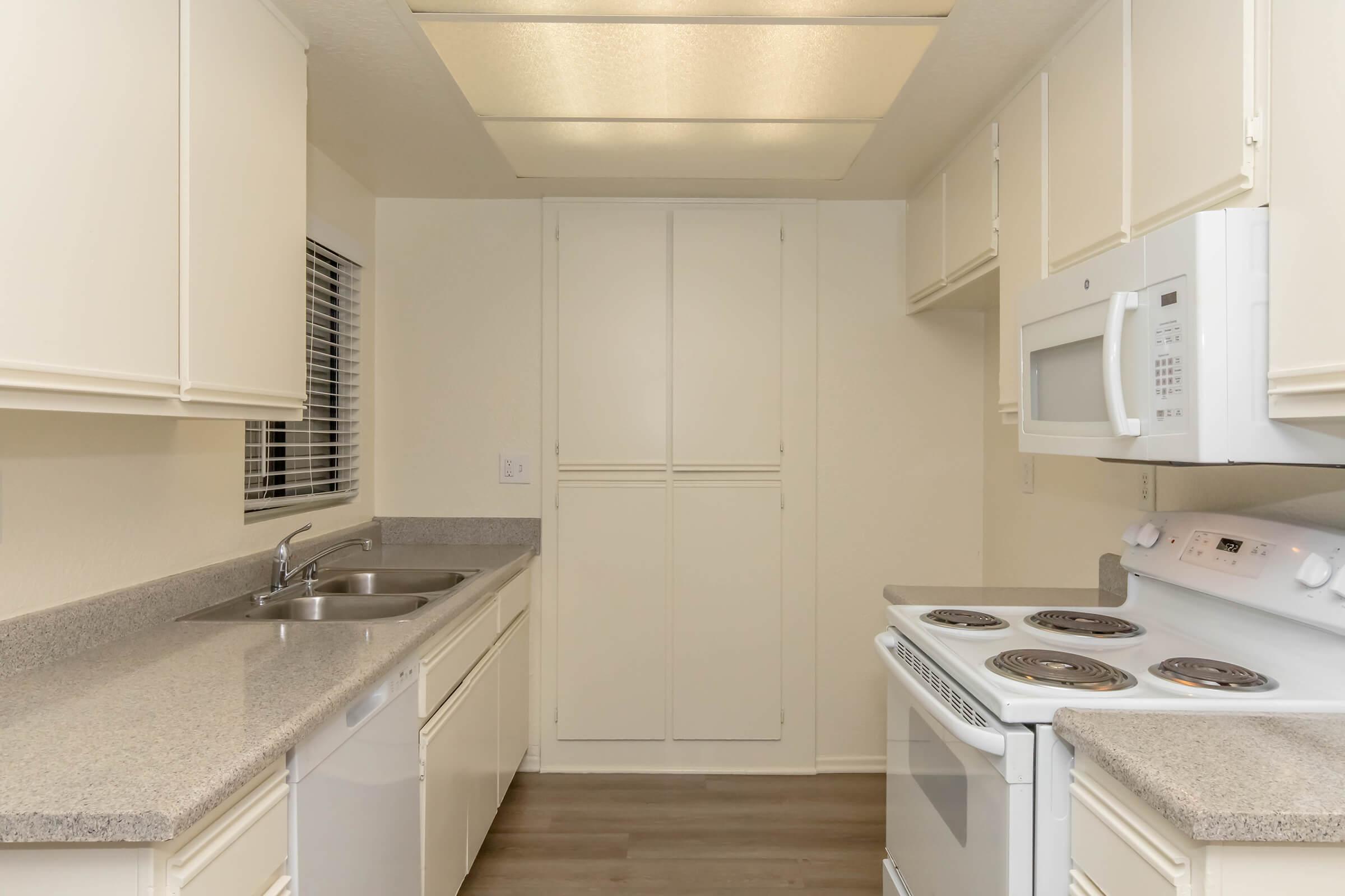 A clean, modern kitchen featuring white cabinets, a double sink, a countertop, a white microwave, and an electric stove with four burners. The space has neutral-colored walls and flooring, with ample natural light coming from a window on the left. Storage cabinets are visible, providing a tidy and functional look.