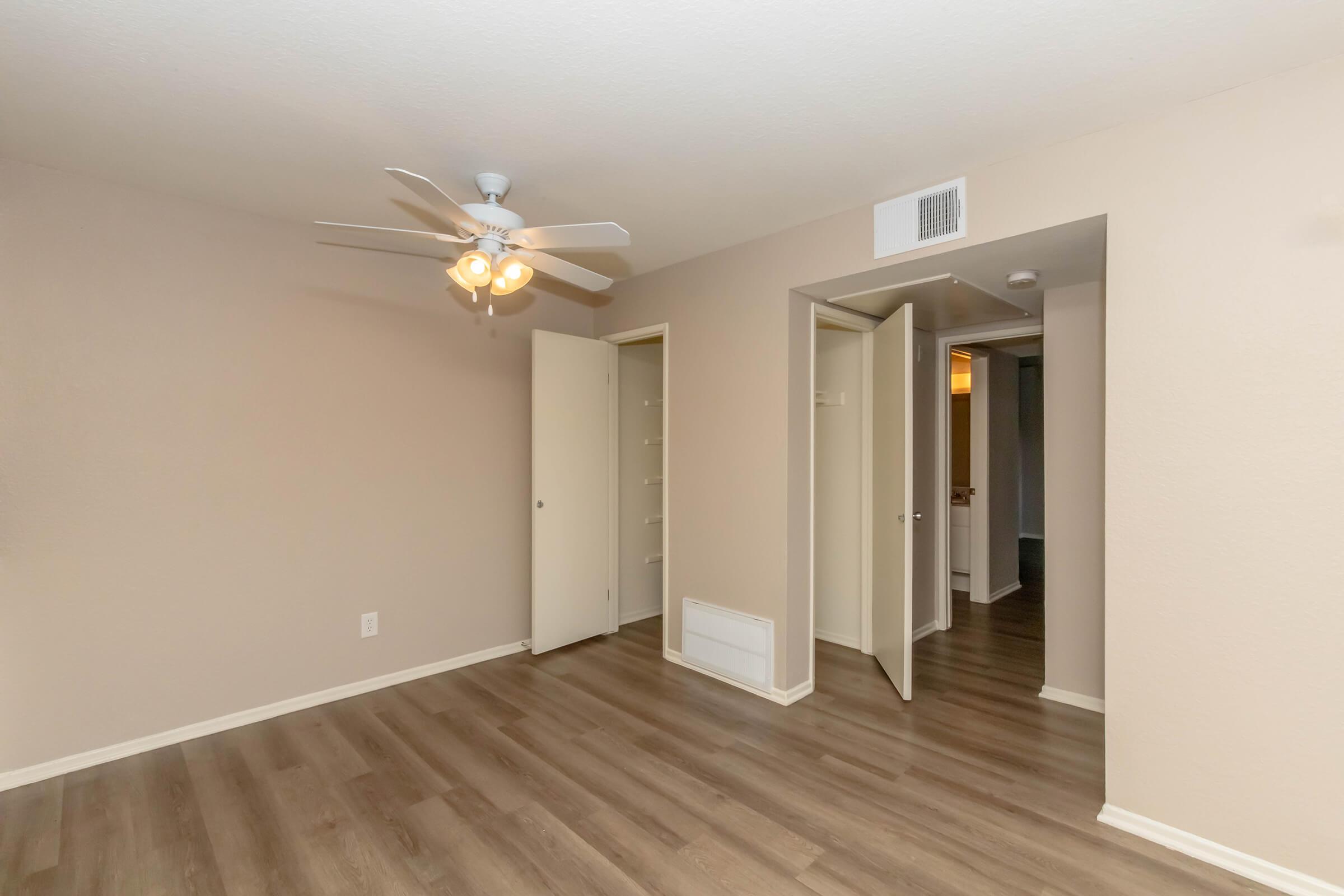 A spacious, light-colored room featuring a ceiling fan, two open closet doors, and a view into a hallway with additional doors. The flooring is a light wood-style laminate, and the walls are painted in a neutral tone, creating a bright and inviting atmosphere.