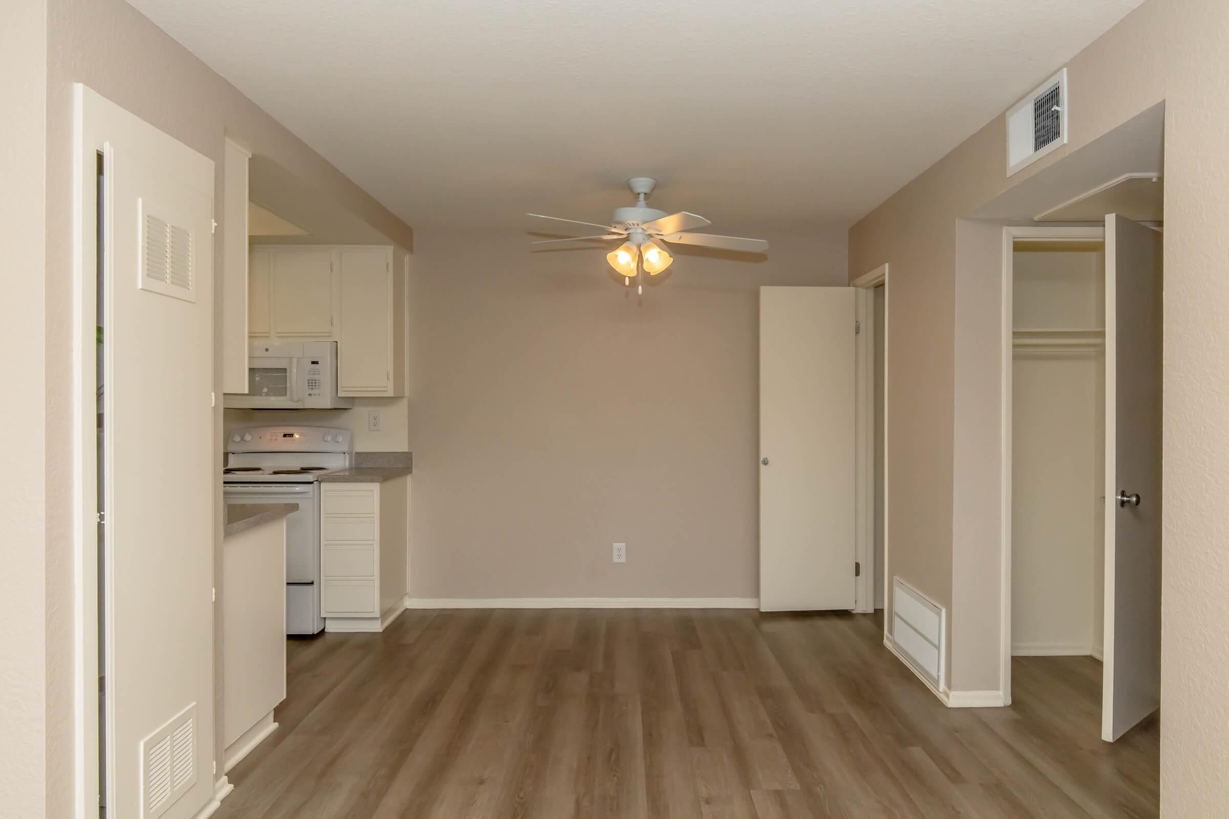 An open living space featuring light-colored walls, a ceiling fan with four lights, and wood-style flooring. To the left, a kitchen area includes white cabinets and appliances. A doorway leads to a closet, and another door opens to a small hallway. The overall atmosphere is bright and spacious.