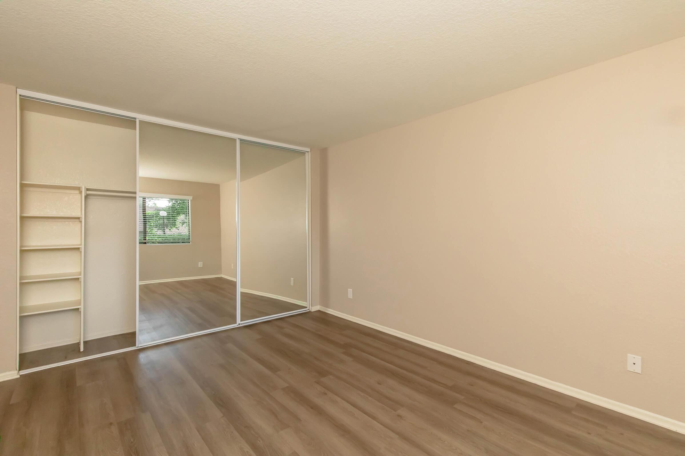 Empty room with light brown walls and laminate flooring. A large sliding mirror closet is on one side, with a small shelf unit beside it. A window is visible in the background, allowing natural light to fill the space. The overall atmosphere is open and inviting, suitable for decoration and personal touches.