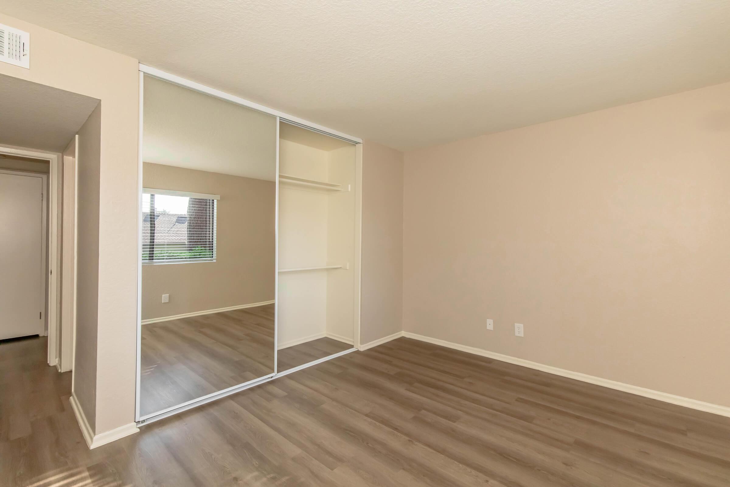 A spacious room with light beige walls and laminate flooring. A mirrored sliding closet door is on one wall, reflecting the neutral tones. A window lets in natural light, providing a view of the outdoors. The overall ambiance is calm and inviting, ideal for a bedroom or living space.