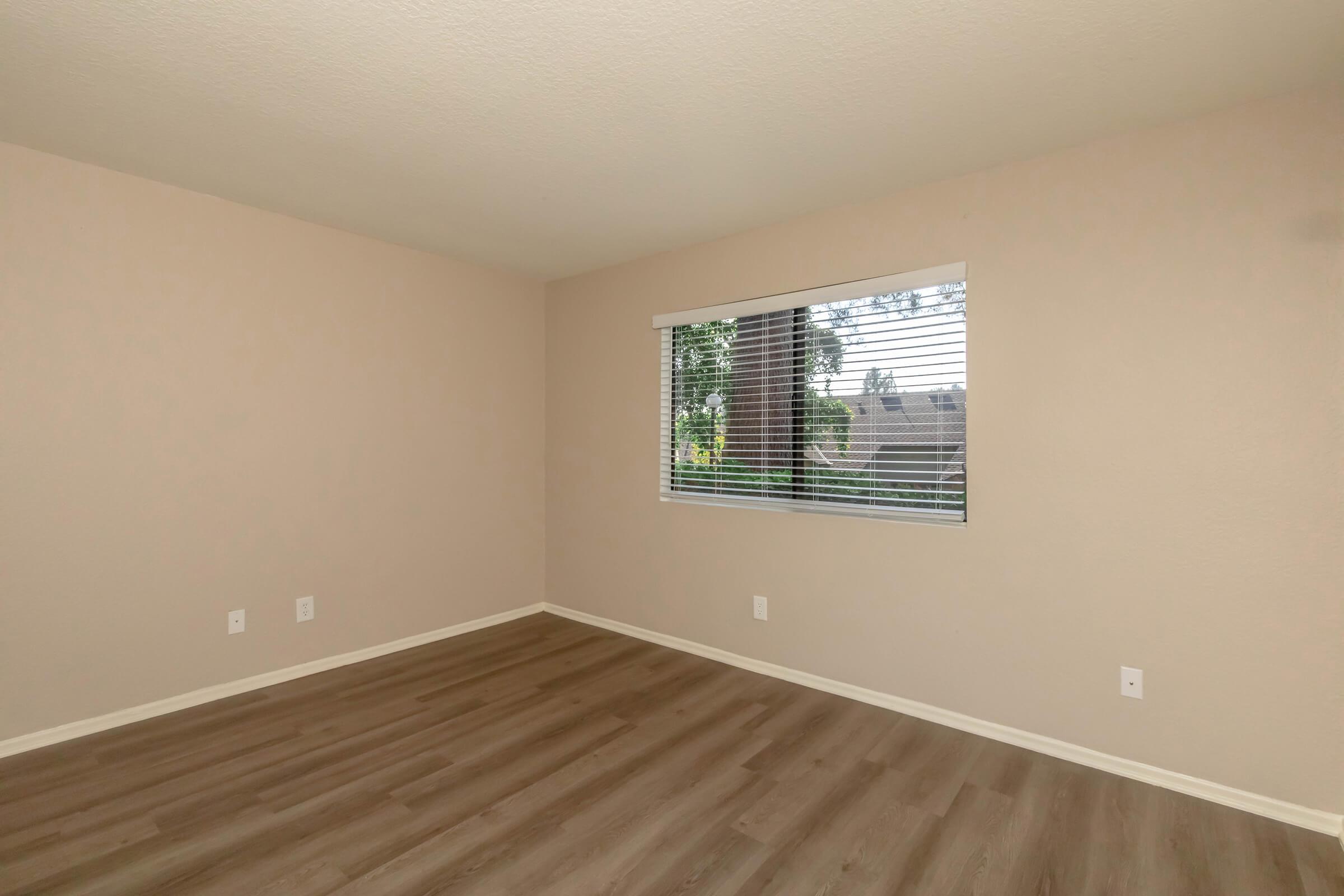 Empty room with light beige walls and laminate flooring. A large window with white blinds allows natural light to enter, offering a view of greenery outside. The room is uncluttered and spacious, creating a bright and open atmosphere.