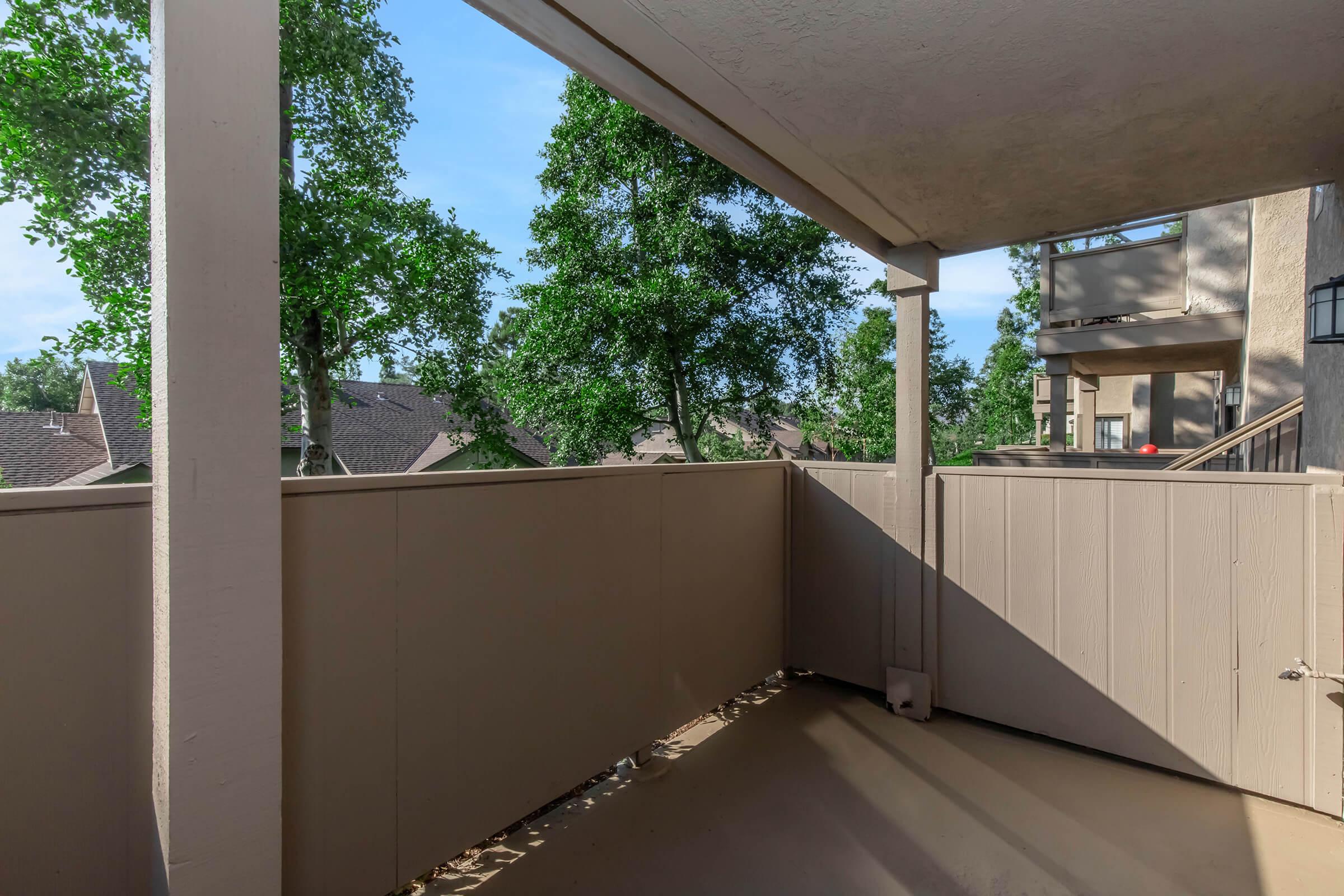 A balcony view featuring a wooden railing and a beige wall. In the background, there are tall trees and rooftops of nearby buildings under a clear blue sky. The area is well-lit, suggesting a sunny day. The floor is plain and free of furniture.