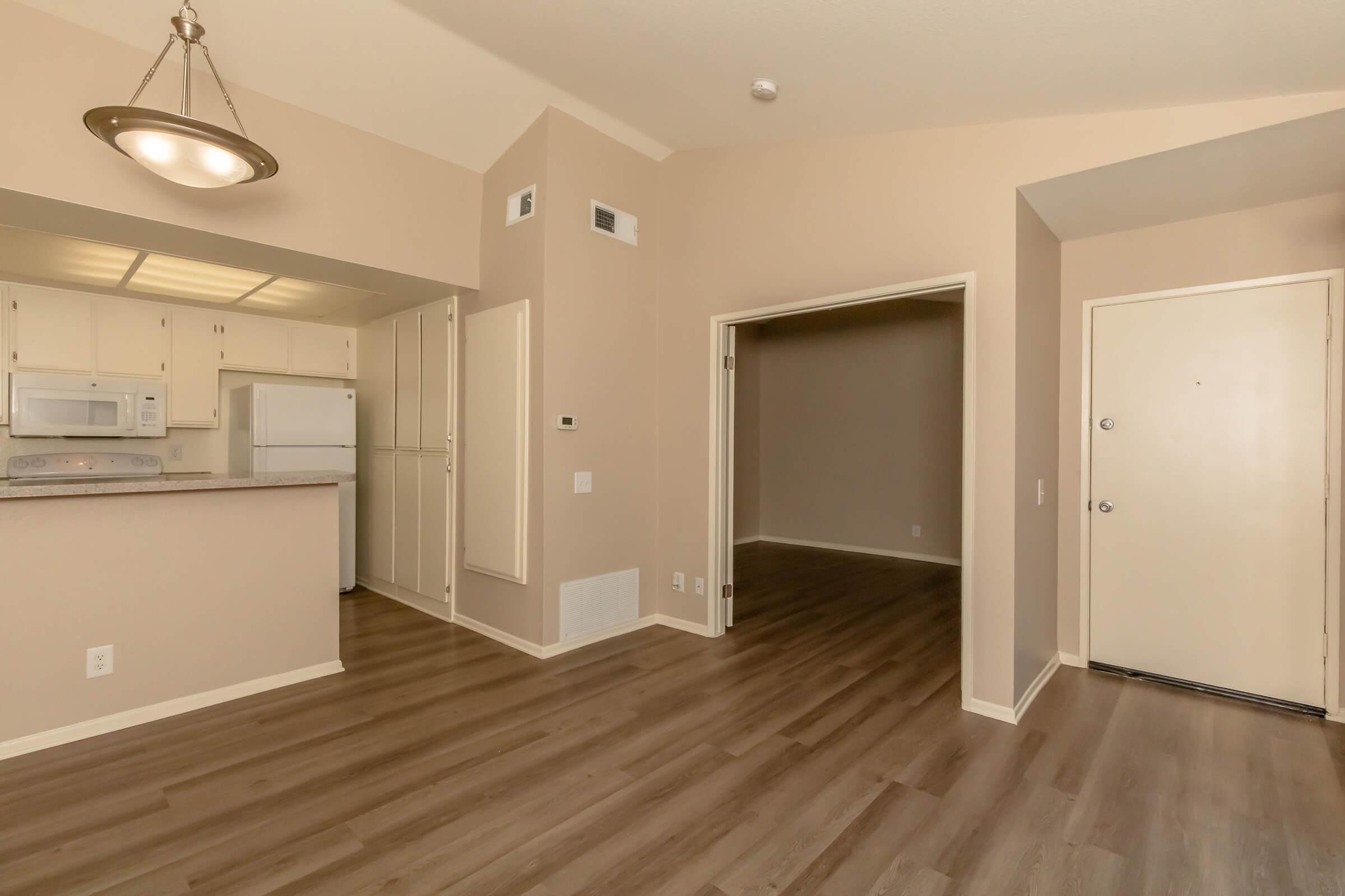 Interior view of a modern kitchen and living area with light-colored walls and hardwood floors. The kitchen features white appliances and cabinets, with a light fixture above. A doorway leads to another room, and the front door is visible on the right side of the image.