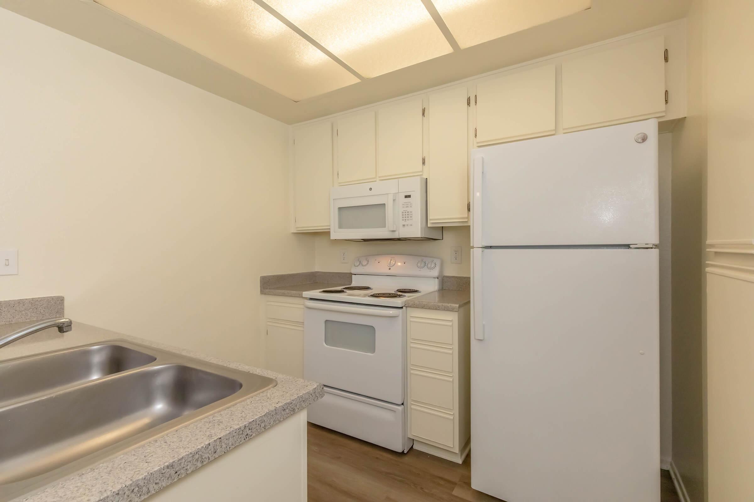 A tidy kitchen featuring white cabinetry, a stove with an oven, a microwave above it, and a refrigerator. The countertops are light gray, and there's a double sink with a simple design. The room has neutral-colored walls and a ceiling with soft lighting.
