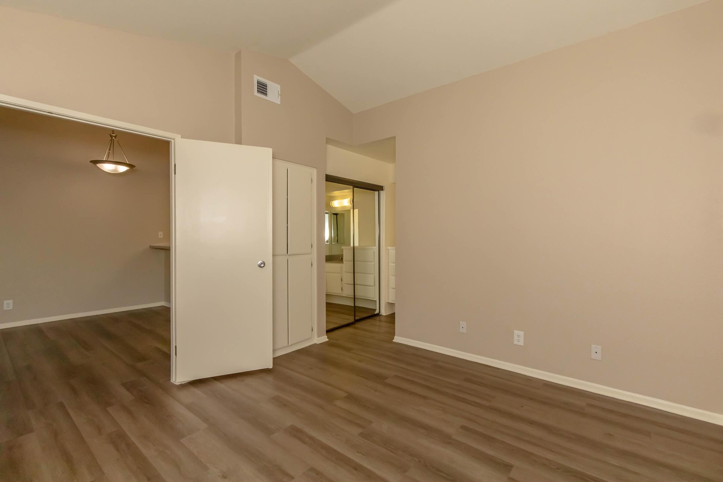 Spacious interior of a light-colored room featuring brown laminate flooring, a doorway leading to another area, and a closet with mirrored doors. Soft natural light illuminates the space, highlighting the neutral wall color and open layout, creating an inviting atmosphere.