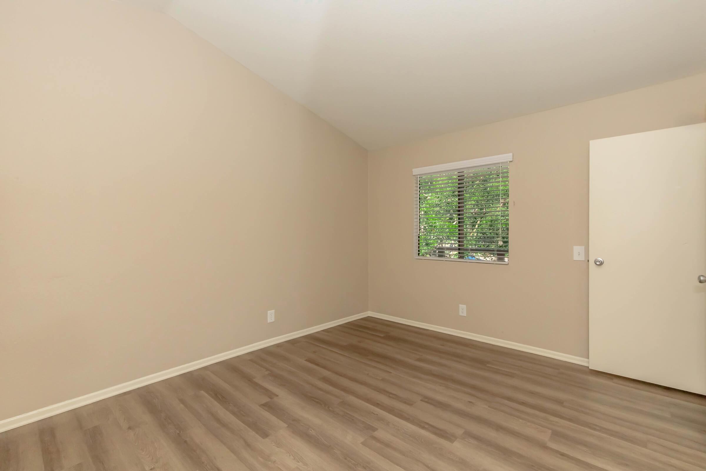 A spacious, empty room with light-colored walls and a high ceiling. There is a window with blinds letting in natural light, and the floor features light wooden laminate. A closed door is visible on the right side of the image. The overall atmosphere is clean and minimalistic, perfect for personal customization.