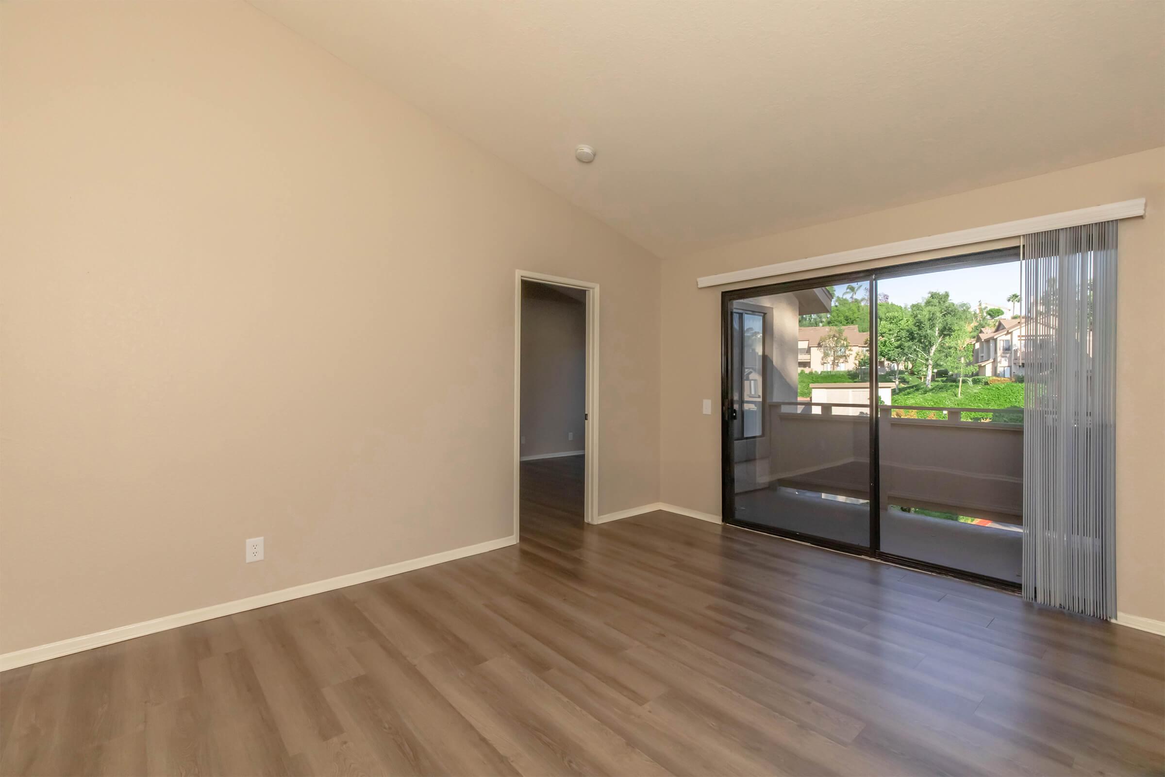 A spacious, lightly colored living area with high ceilings, featuring hardwood flooring and large sliding glass doors that lead to a balcony or outdoor area. A doorway to another room is visible on the left, while the overall ambiance is bright and inviting.