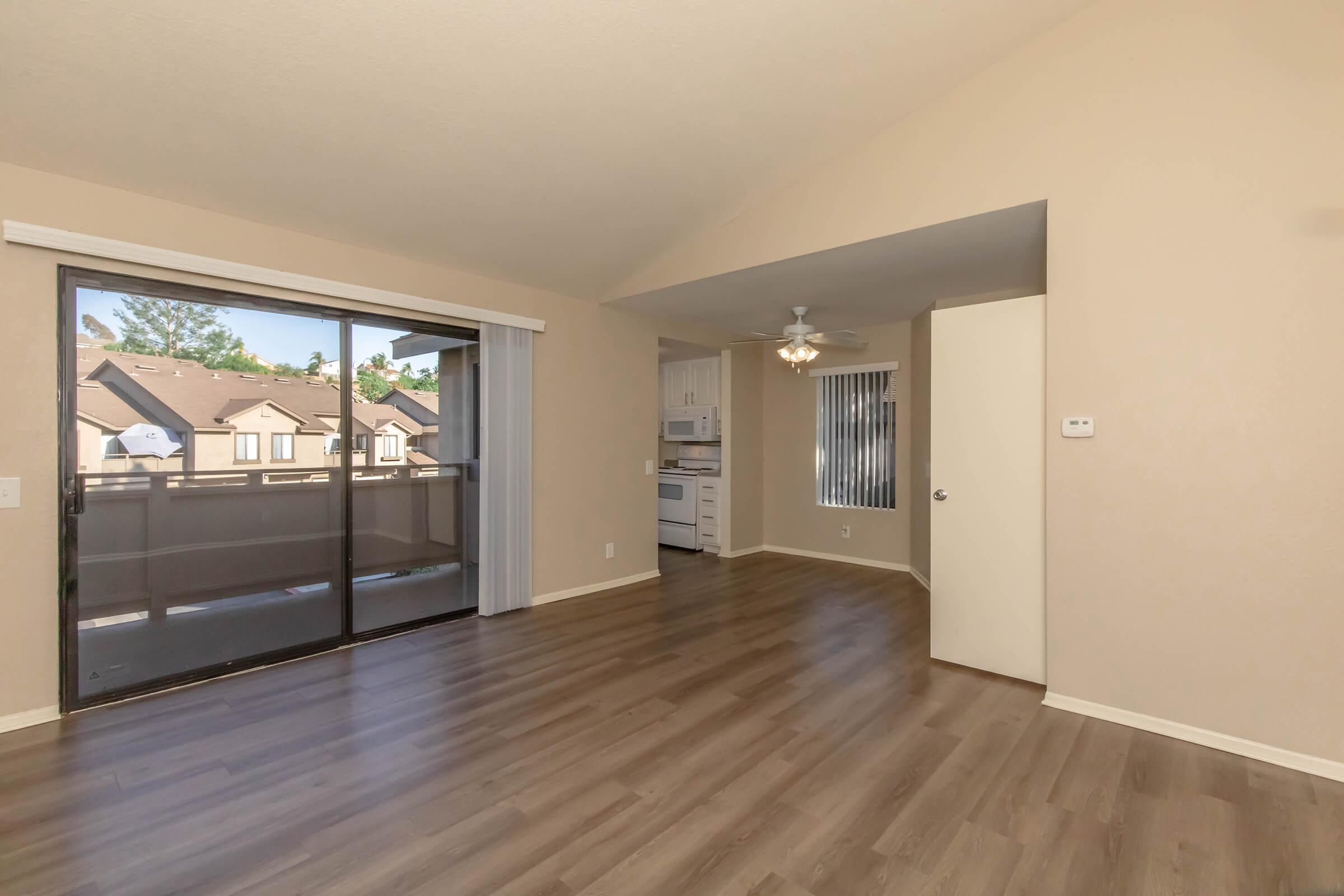 Interior view of a spacious living room featuring light-colored walls and wood-style flooring. Large sliding glass doors provide natural light and open to a balcony. A ceiling fan is visible, and a doorway leads to a kitchen area with white cabinetry. The overall ambiance is bright and inviting.