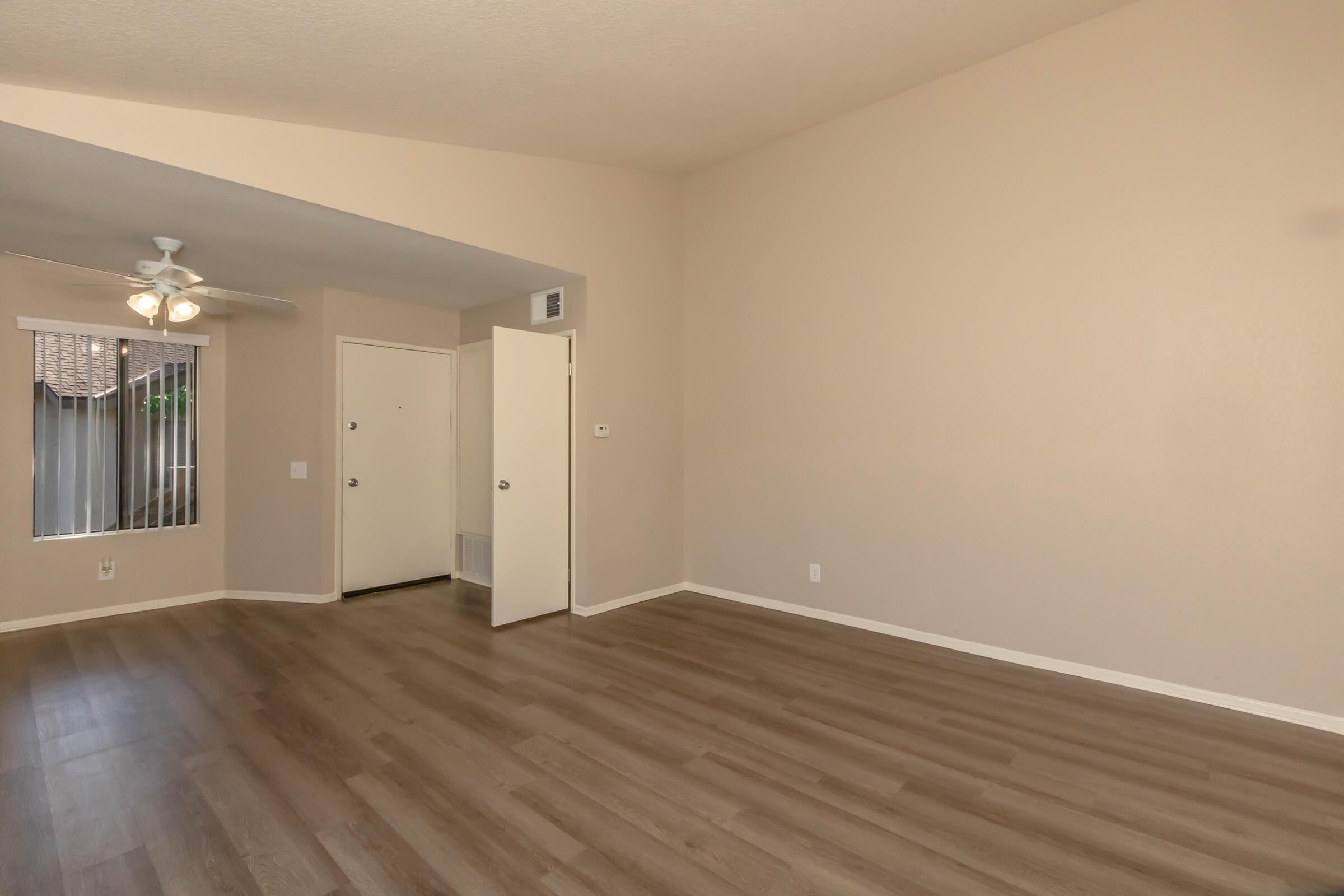 A spacious, light-filled interior of a room with a light-colored wall and wooden flooring. There is a ceiling fan, an entrance door, and large windows with blinds. The overall ambiance is clean and modern, suggesting a cozy living space.