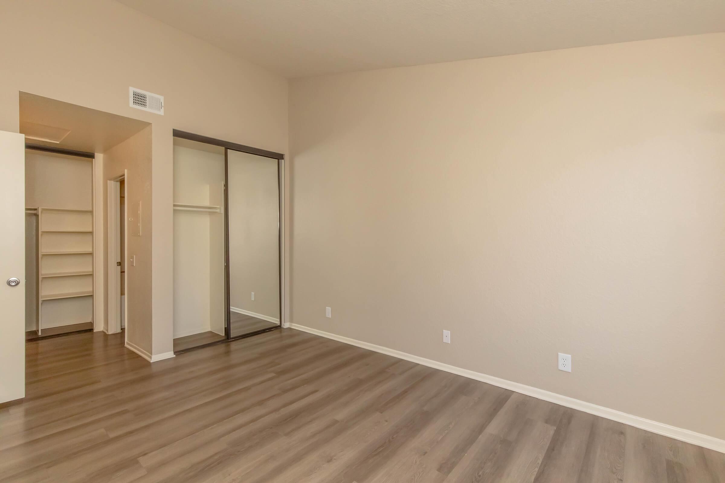 A spacious, empty room with light beige walls and a high ceiling. The floor is light-colored wood laminate. To the left, there is an open doorway leading to a closet with built-in shelves. A mirrored sliding door is visible in the closet area. The overall atmosphere is bright and minimalistic.