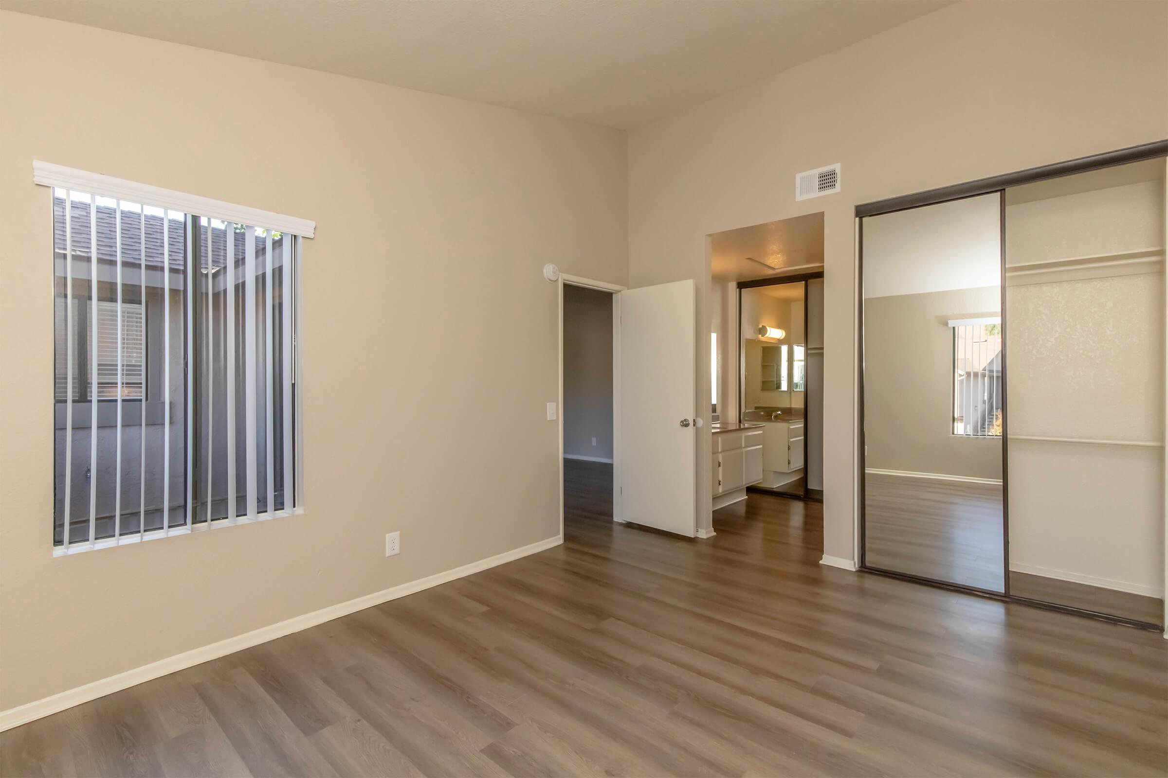 A bright and spacious room with light beige walls and wooden flooring. It features a large window with vertical blinds, open doorways leading to another area, and a mirrored closet. Additional lighting from an adjacent bathroom can be seen in the background, adding to the inviting atmosphere.