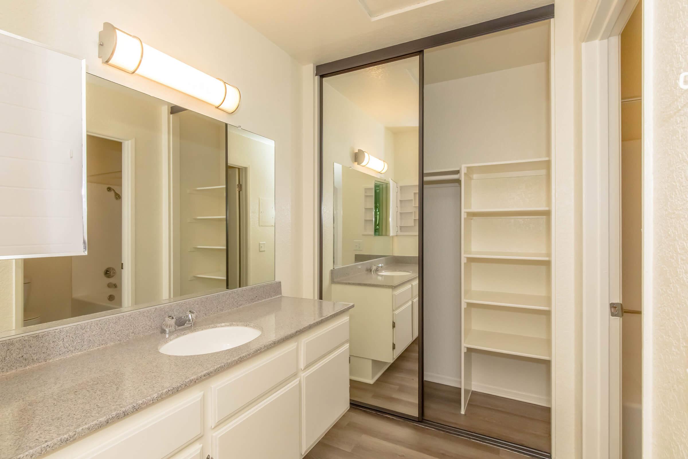 Bright, modern bathroom featuring a large mirror, double sink vanity with gray countertops, and ample cabinet space. A mirrored closet reflects the light, and there are shelves for storage. The walls are painted a light color, and the flooring is a wood-like laminate.