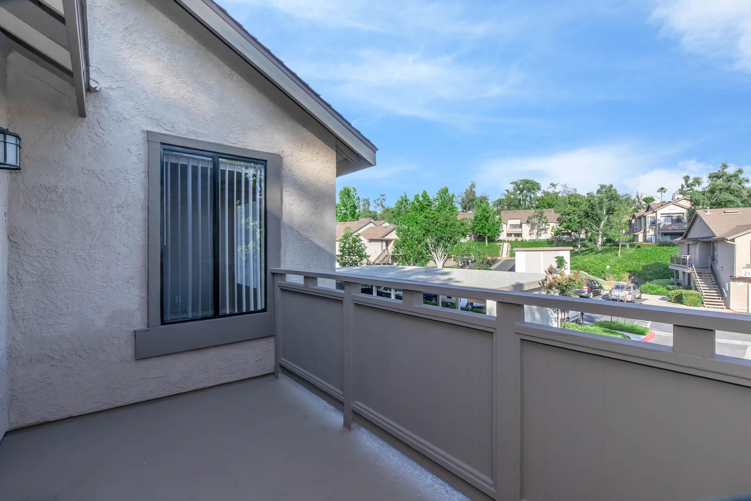 A clean balcony with a railing overlooking a residential area. The balcony features a sliding glass door with vertical blinds and is surrounded by trees and buildings in the distance, under a clear blue sky. The area appears well-maintained and inviting.