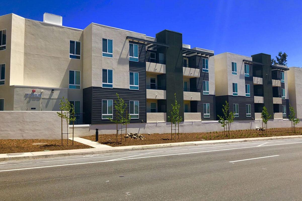 A modern apartment building featuring a combination of light and dark exterior colors, with multiple balconies, large windows, and landscaping in front.