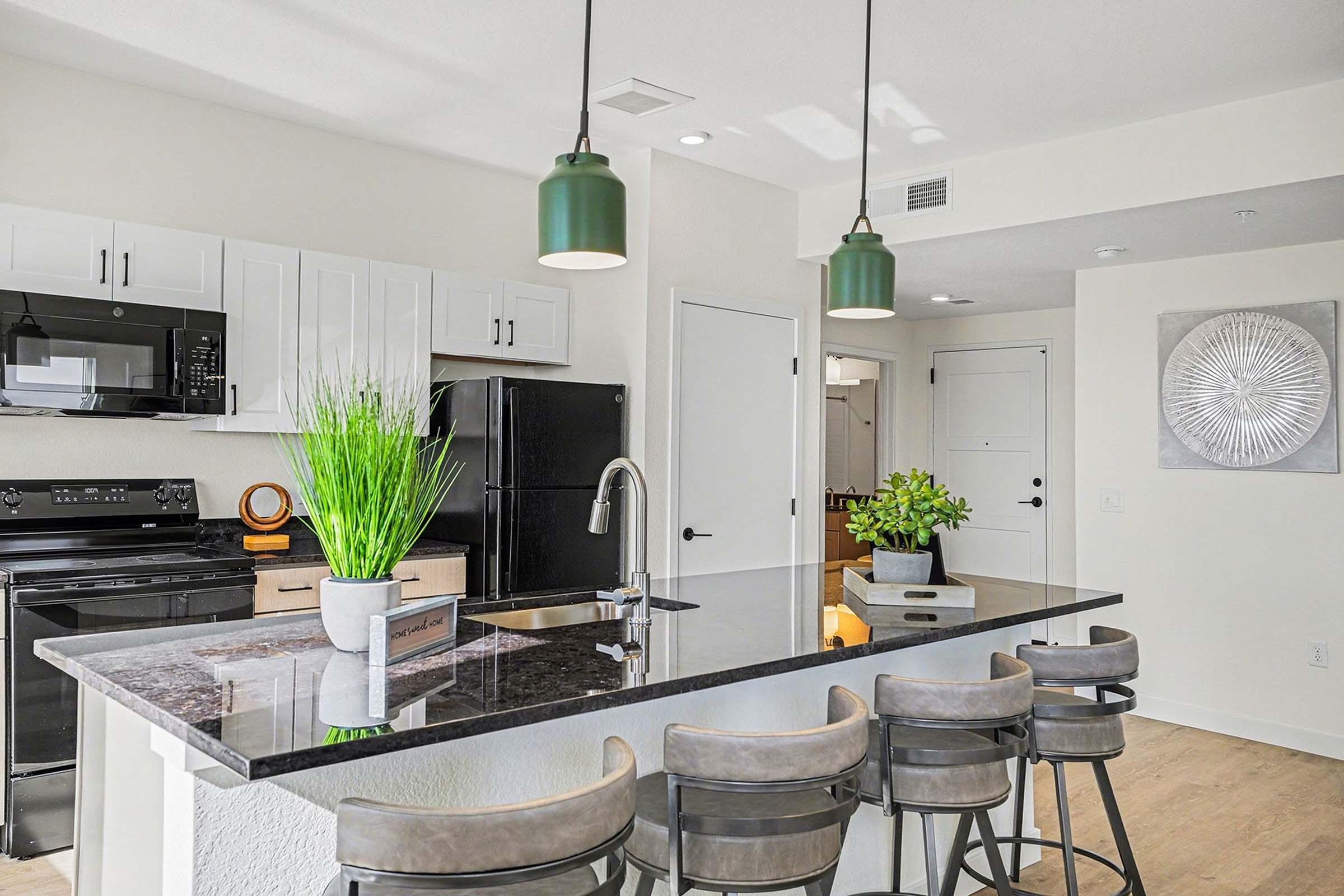 Modern kitchen featuring a large island with seating for four, black countertops, and white cabinetry. The space includes a microwave and a black refrigerator, with green pendant lights hanging above the island. Decorative plants are placed on the counter, enhancing the contemporary design.