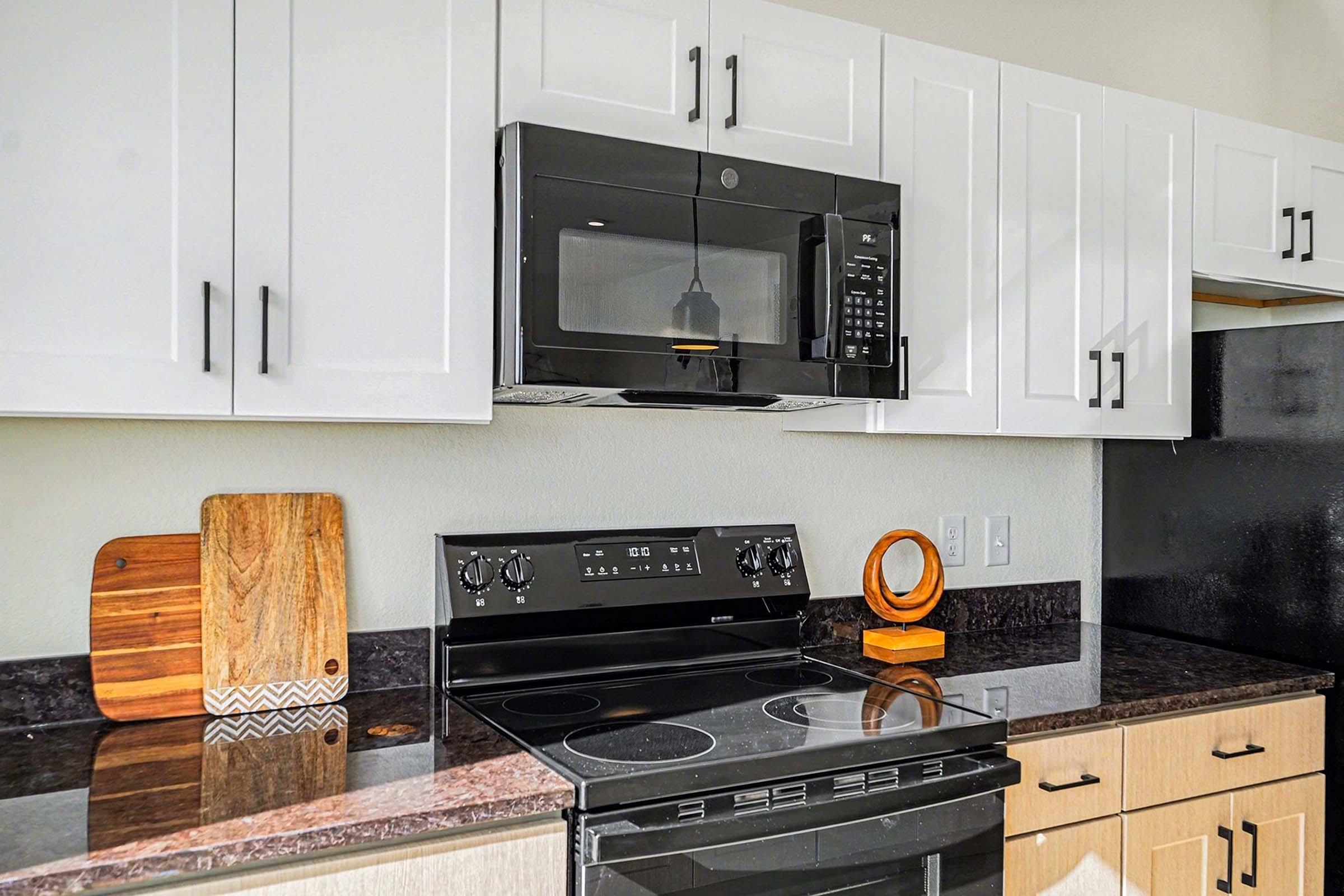 A modern kitchen featuring a black microwave mounted above a sleek black electric stove. The countertop is dark with a wooden cutting board and a decorative wooden circular piece. The cabinetry above is white with simple black handles, creating a contemporary look.