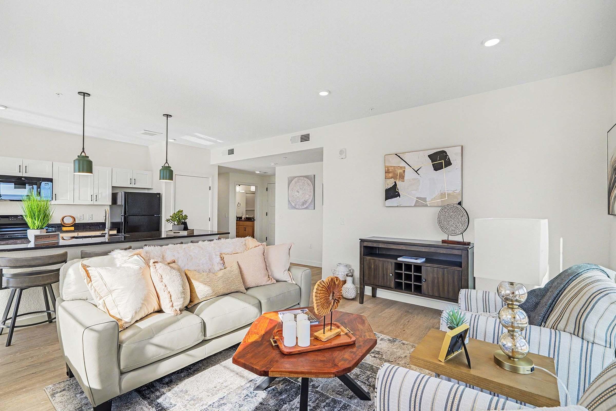 Modern living room featuring a light-colored leather sofa with decorative pillows, a wooden coffee table, and stylish decor. In the background, there is a kitchen area with white cabinets and dark appliances. Natural light fills the space, creating a bright and inviting atmosphere.