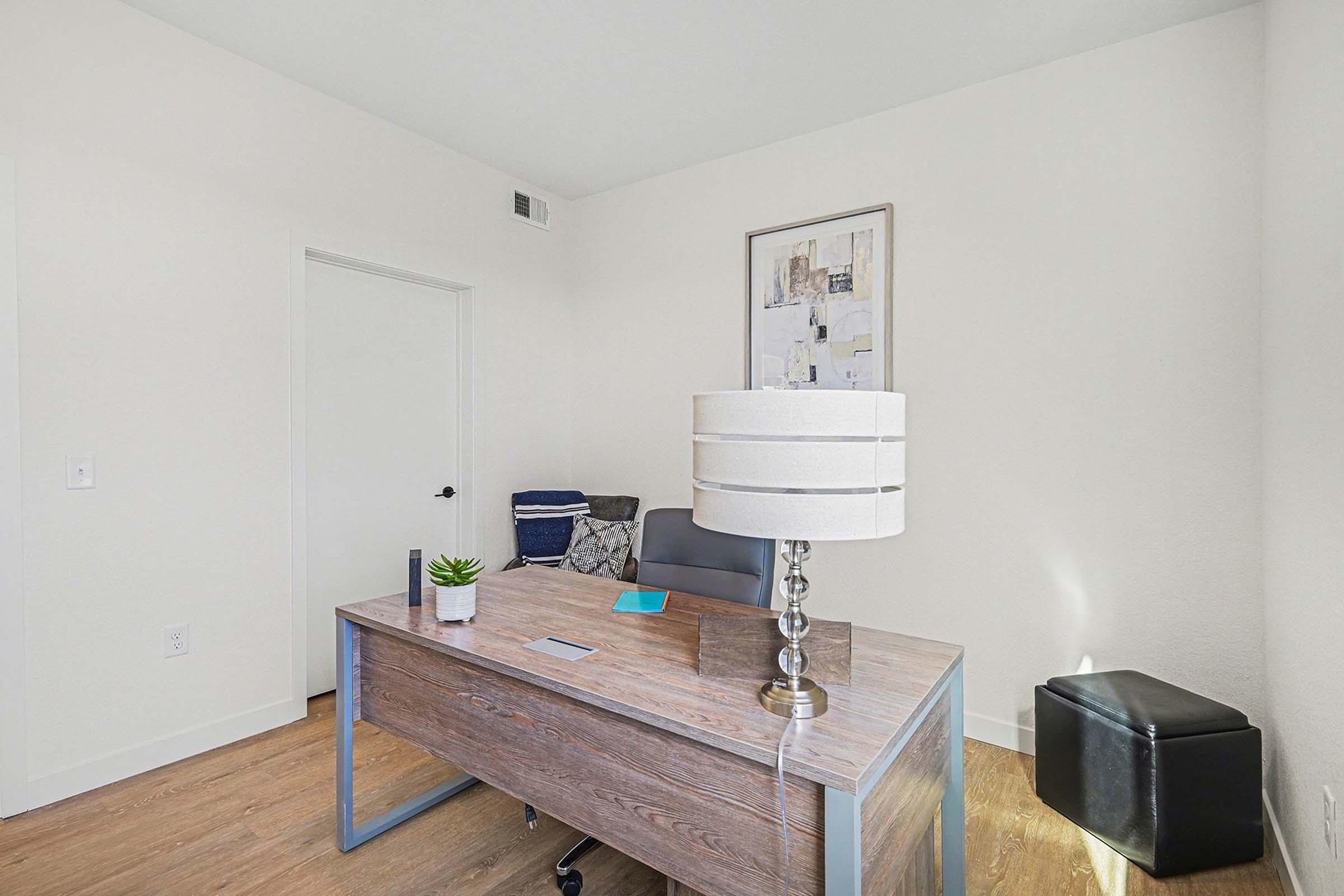 A modern office space featuring a wooden desk with a lamp, a black chair, and a small plant. The room has light-colored walls and wooden flooring. A door is visible, along with a black storage ottoman in the corner and a framed artwork on the wall. Soft natural light illuminates the area.