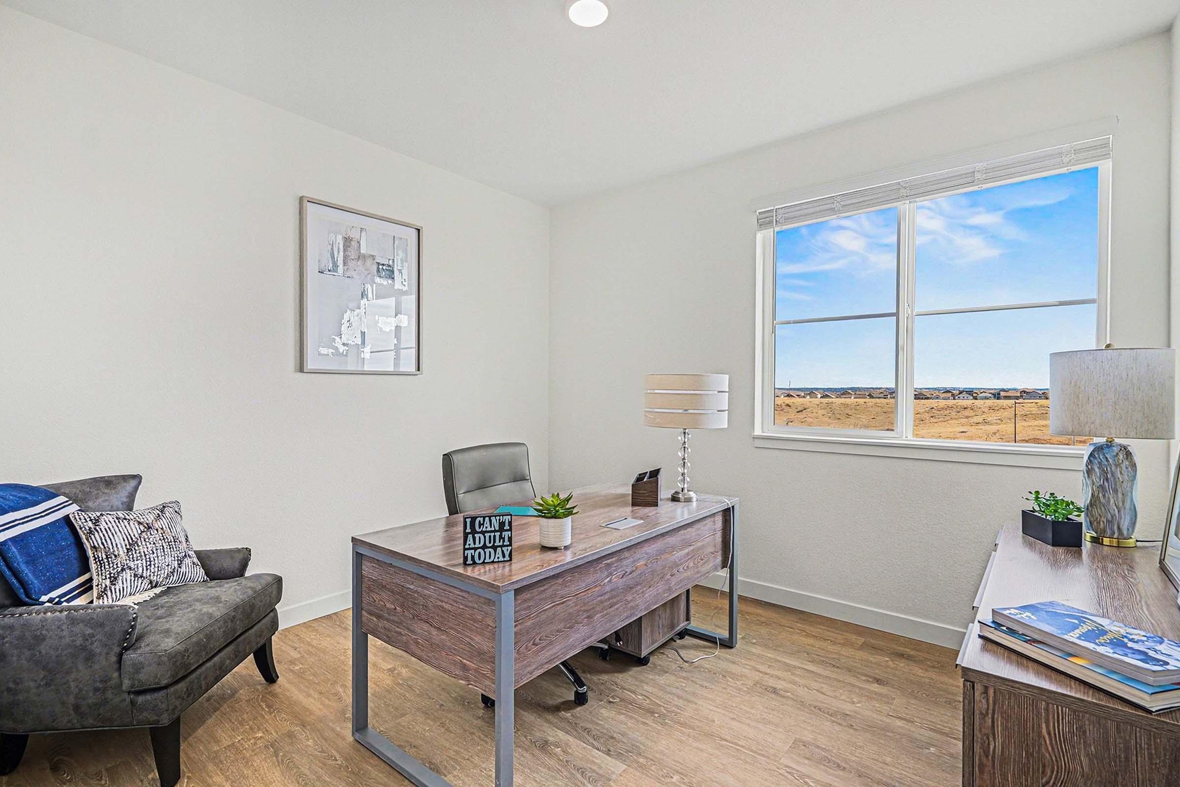 Modern home office featuring a wooden desk with a lamp and decorative items, a comfortable armchair, and a framed picture on the wall. A large window allows natural light and offers a view of a clear sky and distant landscape. The space has a minimalist design with light-colored walls and wooden flooring.