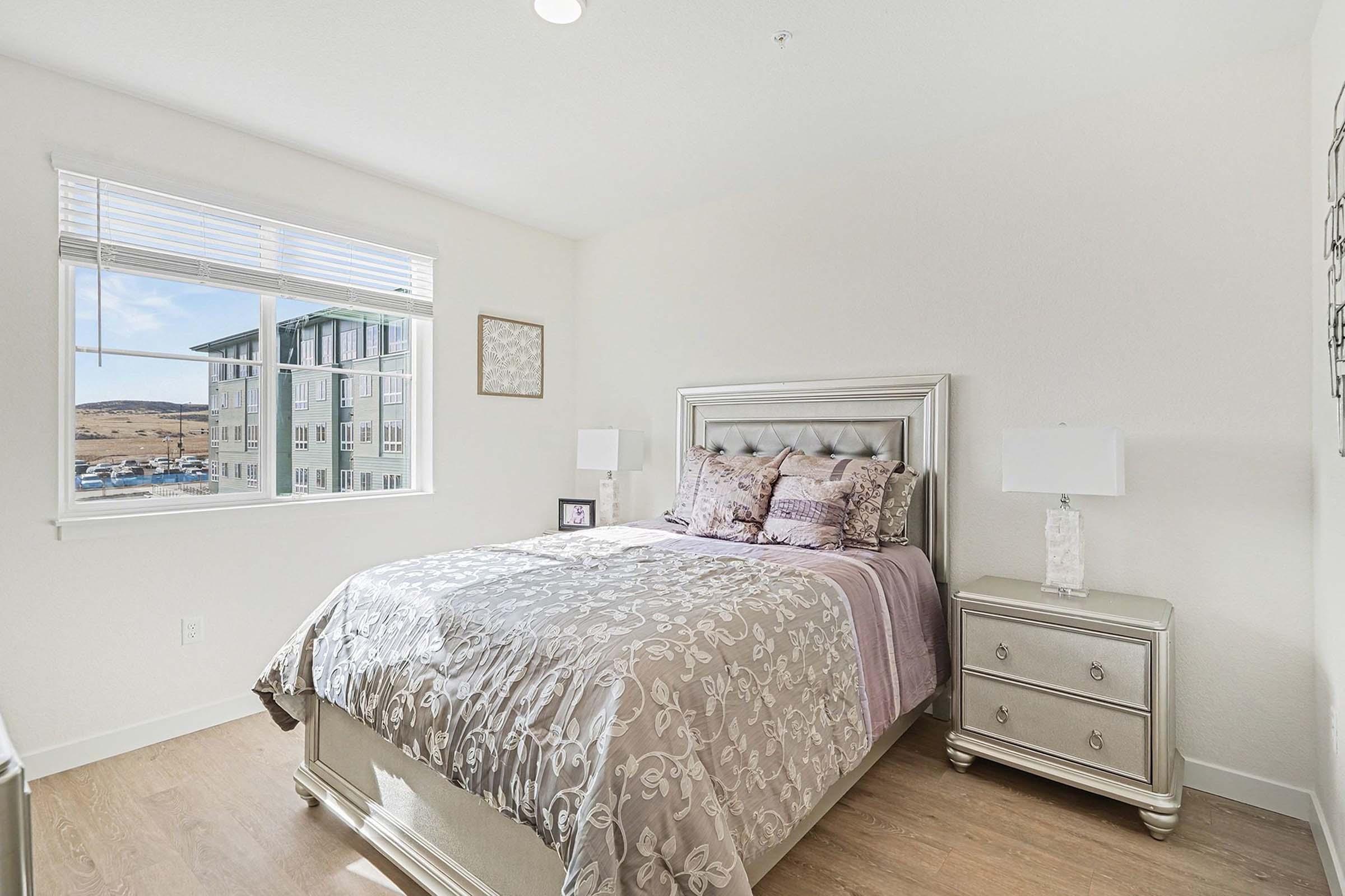 A well-decorated bedroom featuring a queen-sized bed with a silver upholstered headboard, elegant bedding in shades of purple, a stylish bedside table with a lamp, and a window allowing natural light. Modern decor and neutral walls enhance the cozy atmosphere.