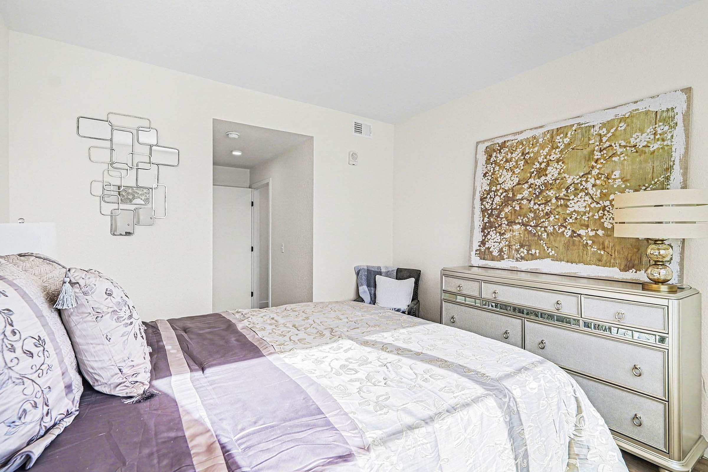 A cozy bedroom featuring a neatly made bed with decorative pillows, light-colored walls, and a stylish dresser. A large piece of framed art with floral details is mounted above the dresser, and a mirrored wall decor piece adds a touch of elegance. Soft lighting creates a warm atmosphere.