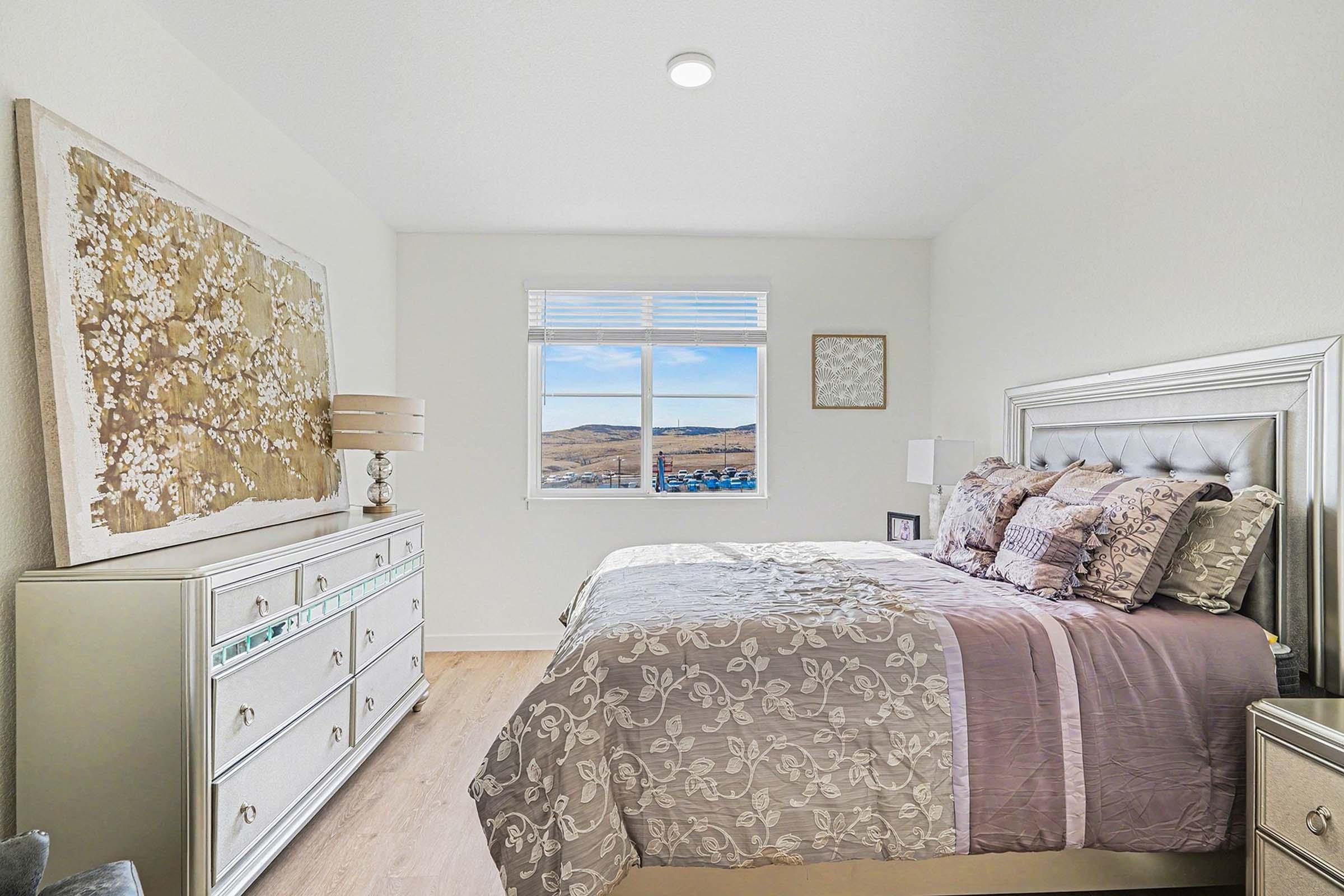 A cozy bedroom featuring a large bed with decorative pillows, a stylish lamp on a nightstand, and a beige and white color palette. A tall dresser stands against one wall, and a window offers a view of rolling hills outside. Soft lighting creates a warm and inviting atmosphere.