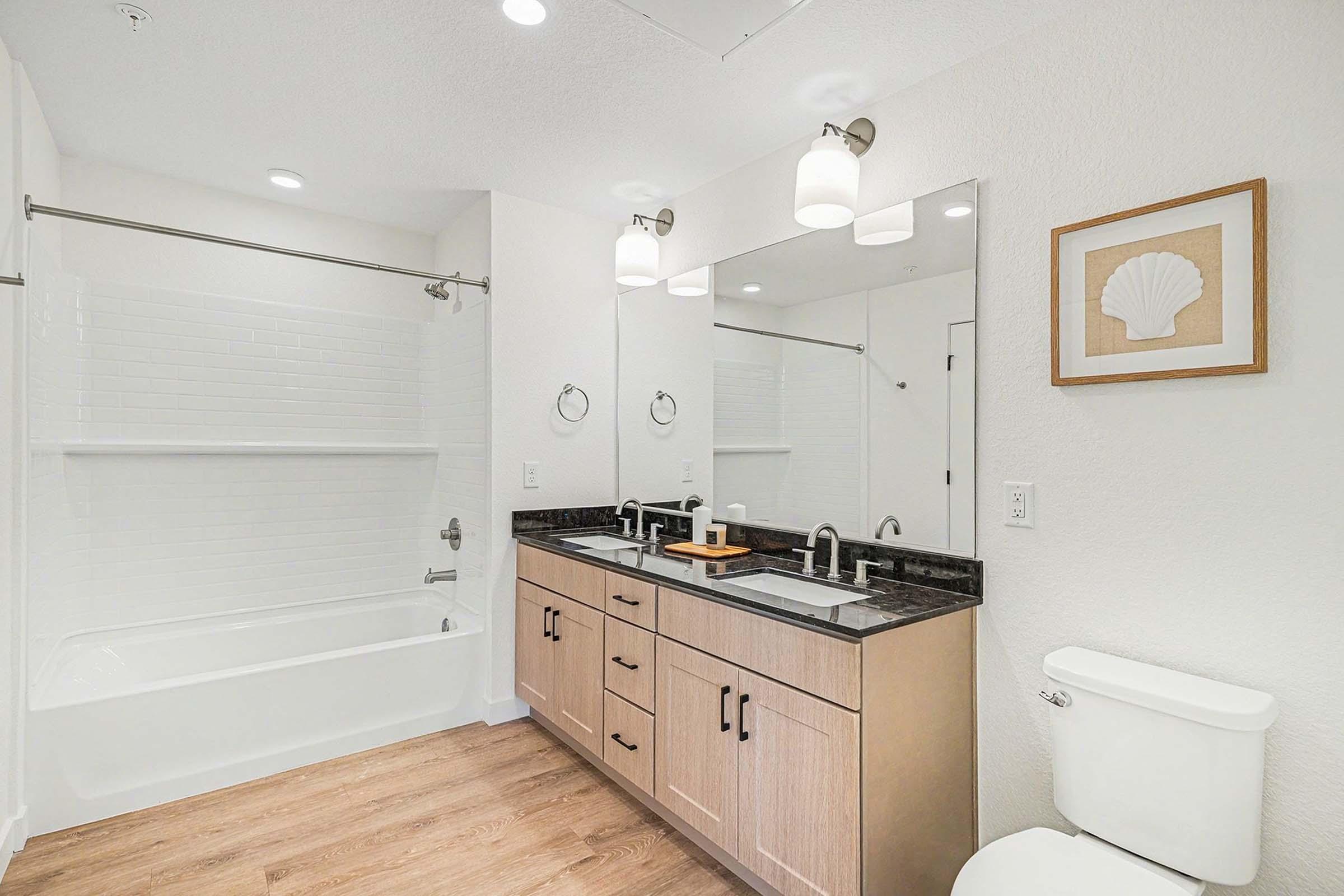 A modern bathroom featuring a white tub and shower combination, dual sinks with a dark countertop, wooden cabinetry, and a large mirror. The walls are painted white, and the flooring is a light wood. A decorative shell print is framed on the wall, and soft lighting fixtures illuminate the space.